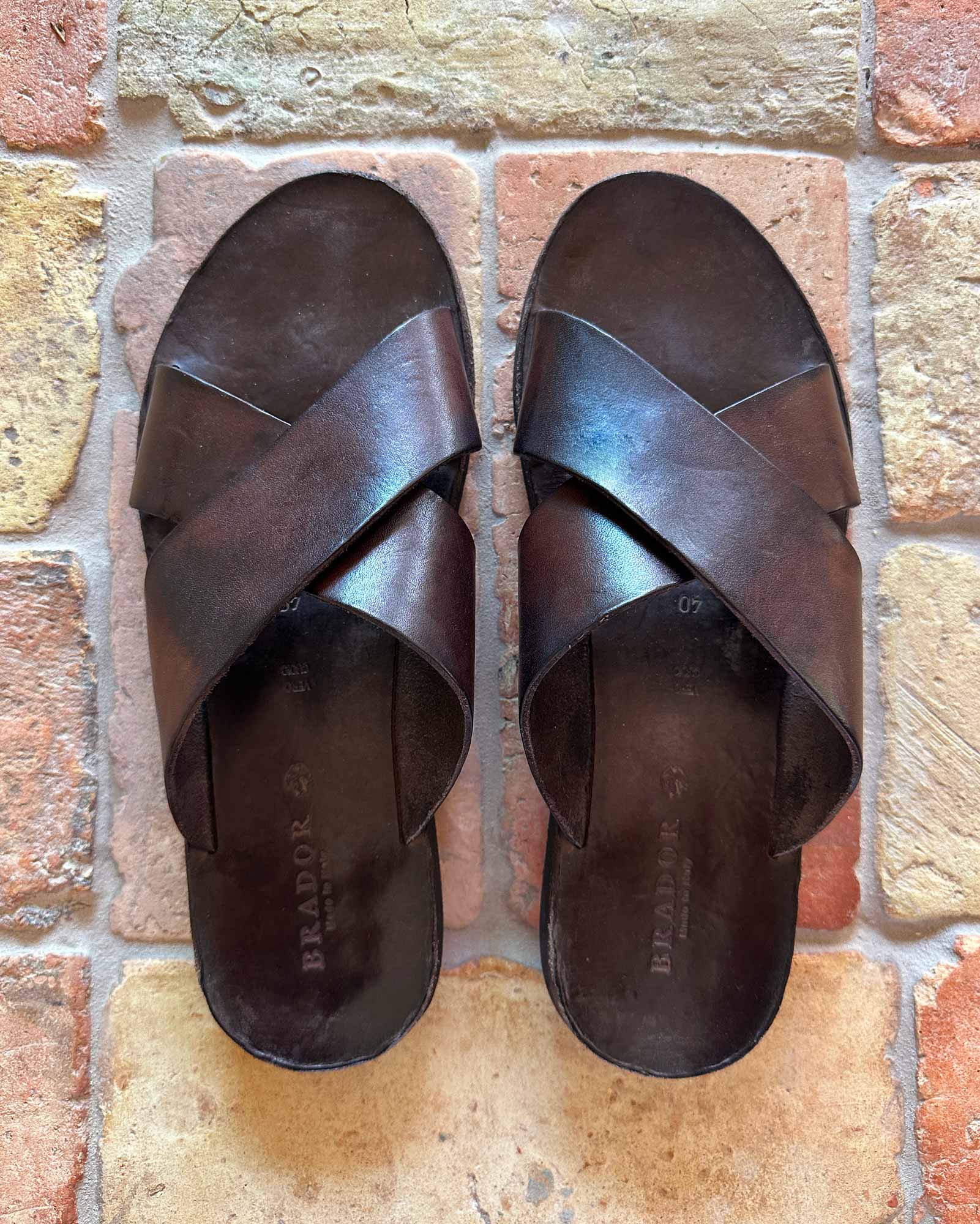 The Dark Brown Leather Infinity Sandal by Pyne and Smith Clothiers, featuring crisscross leather straps, handmade by Italian artisans, is shown side by side on a textured multicolored brick floor.