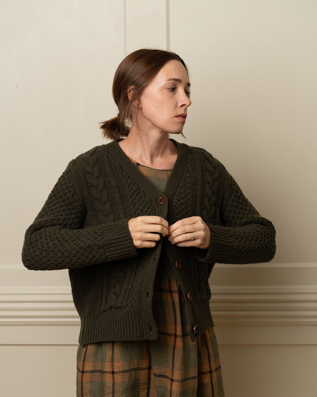 A woman with brown hair in a low ponytail buttons a Pyne and Smith Clothiers Merino Wool Cable Knit Cardigan with Pockets in Brunswick Green over a plaid dress, standing indoors against a light wall and looking to the side.