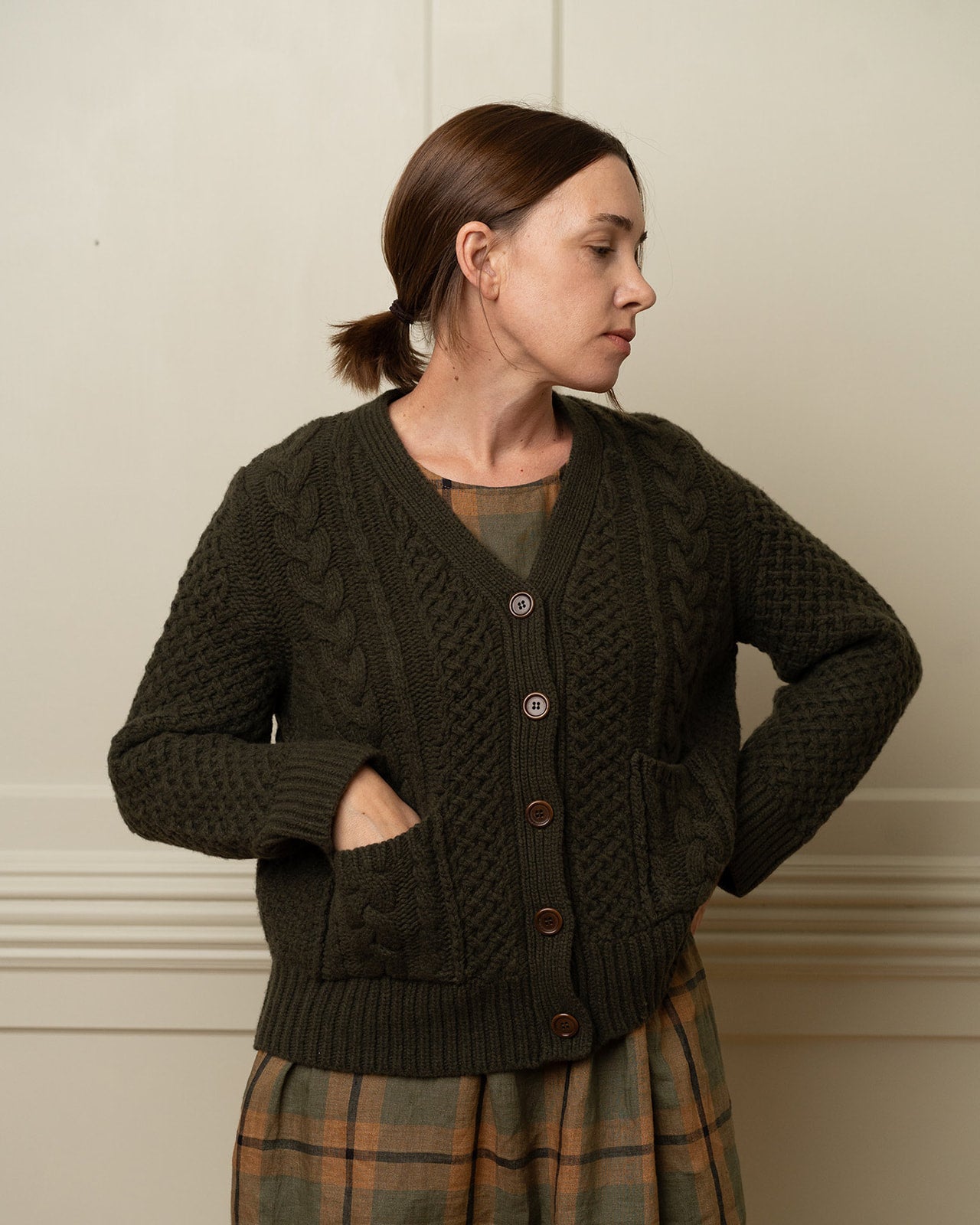 A woman with brown hair pulled back wears the Pyne and Smith Clothiers Merino Wool Cable Knit Cardigan with Pockets in Brunswick Green over a plaid dress, standing against a cream wall, hands on her hips and looking to her left.