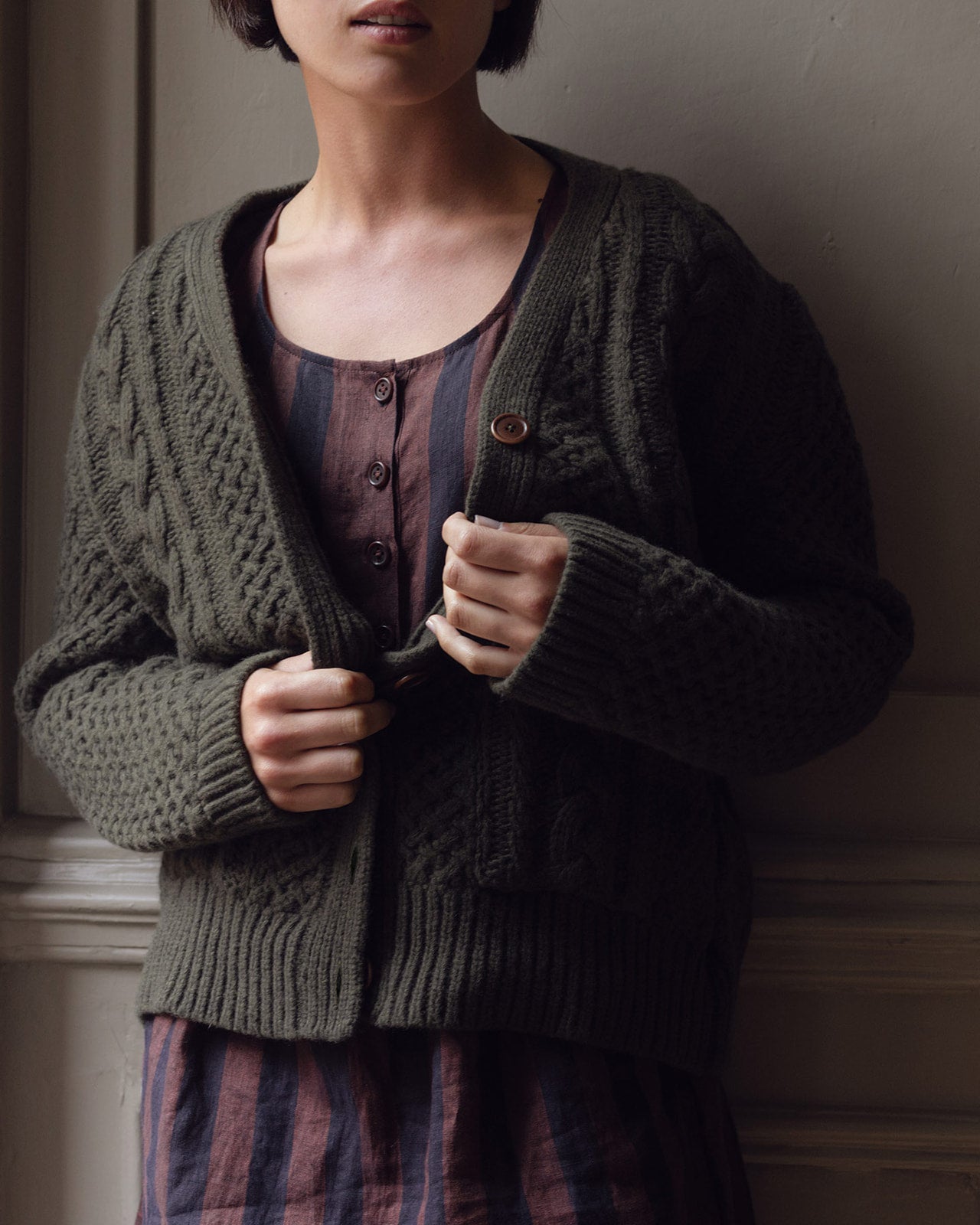 A person partially buttons the Pyne and Smith Clothiers Merino Wool Cable Knit Cardigan with Pockets in Brunswick Green over a brown and purple striped dress indoors, their face partly out of frame.