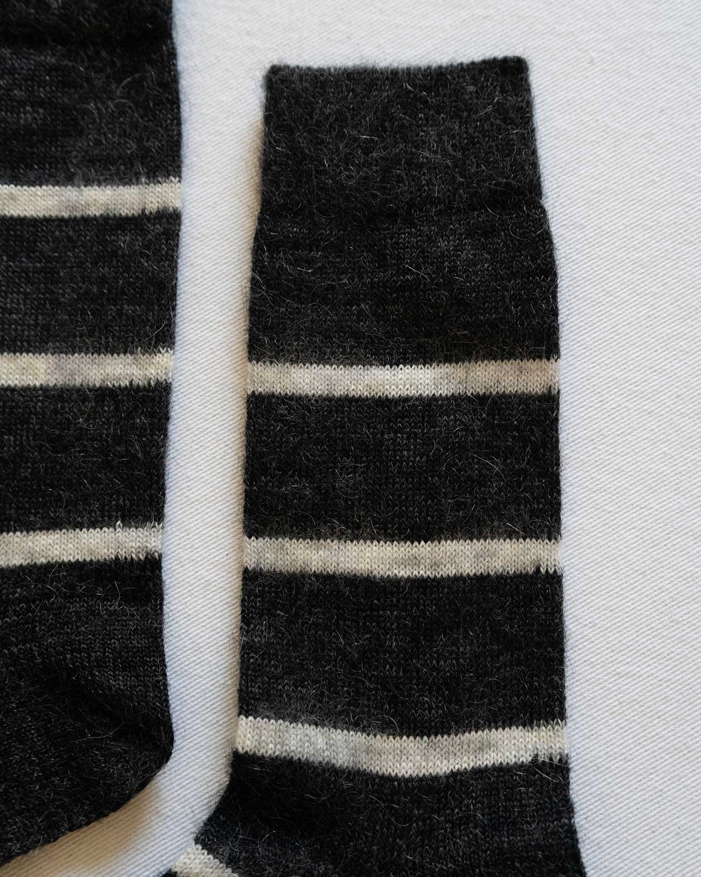 A close-up of Pyne and Smith Clothiers Charcoal Stripe Mohair Wool Socks shows two black, lightweight socks with white horizontal stripes laid flat, displaying only the leg and top sections on a light surface.