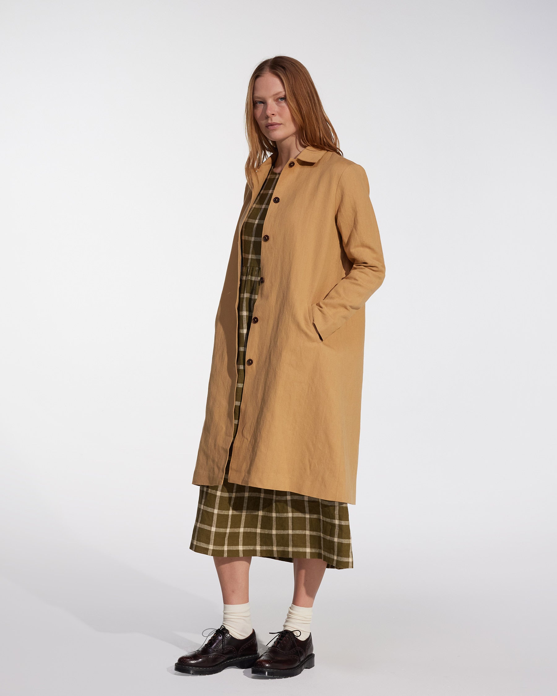A woman with long, straight hair stands against a white background, wearing Pyne and Smith Clothiers' Coat No.11 in Lark Tan Cotton Linen Canvas over a green plaid dress, paired with white socks and dark brown shoes, hands in her pockets.