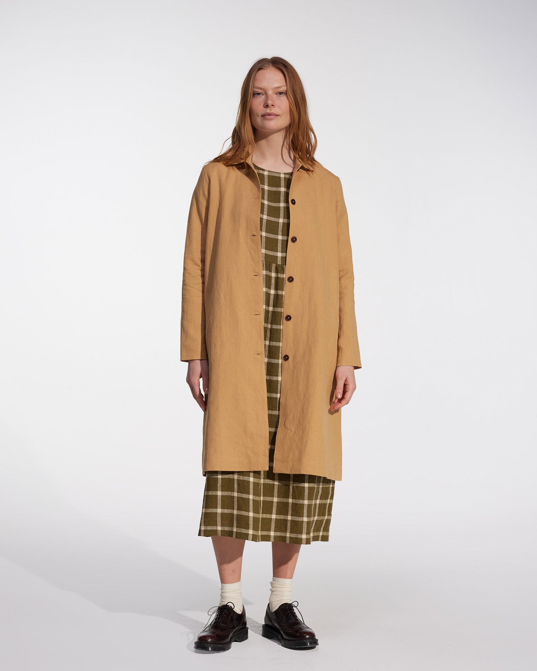 A person stands against a plain white background wearing the Pyne and Smith Clothiers Coat No.11 in Lark Tan Cotton Linen Canvas over a green plaid dress, paired with white socks and black shoes.