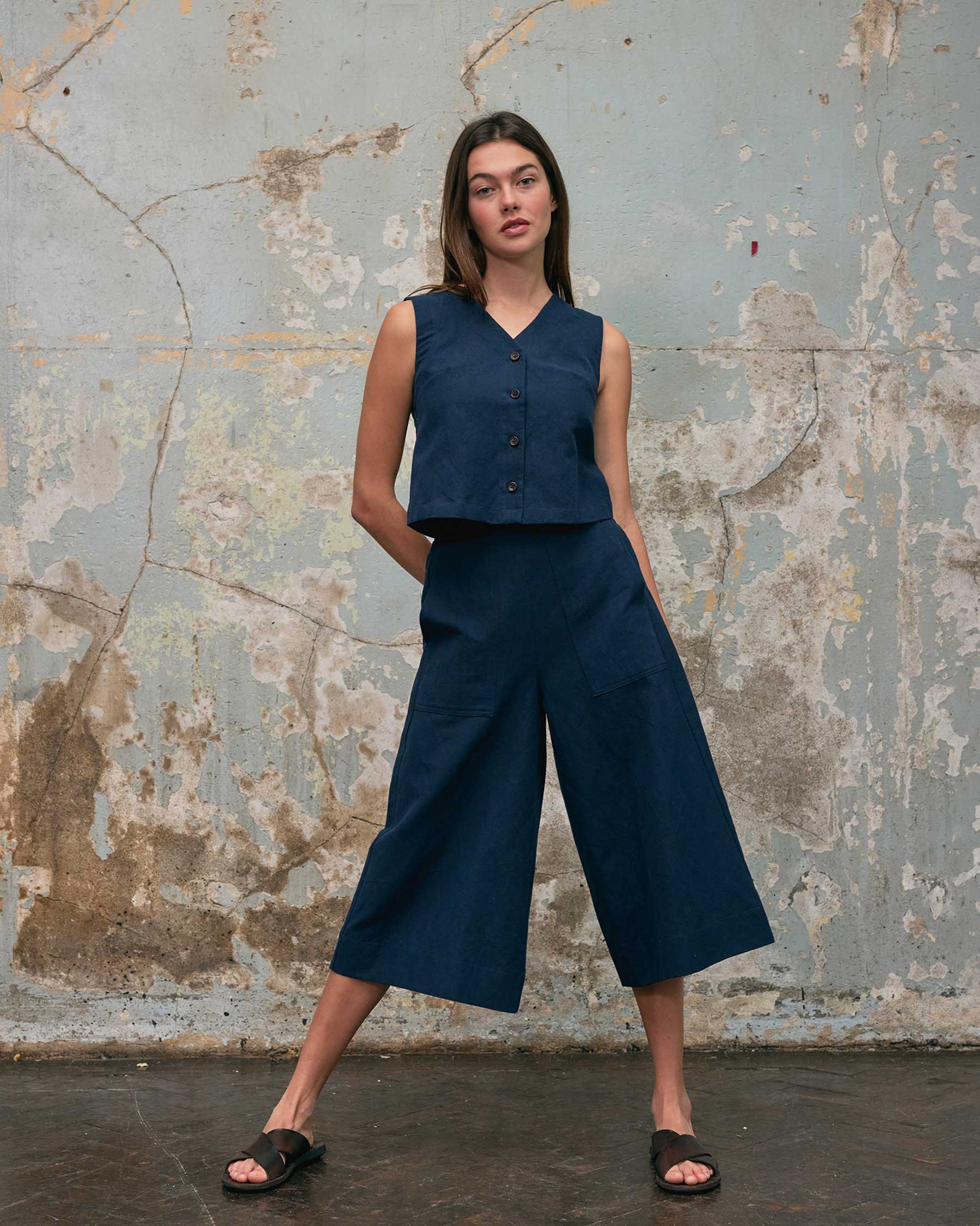A woman stands by a textured wall wearing Pyne and Smith Clothiers' Canvas Cropped Trousers No.17 in Ink Blue, featuring a high-rise waist and wide legs in cotton-linen canvas, paired with a sleeveless top and black sandals. One hand is in her pocket.