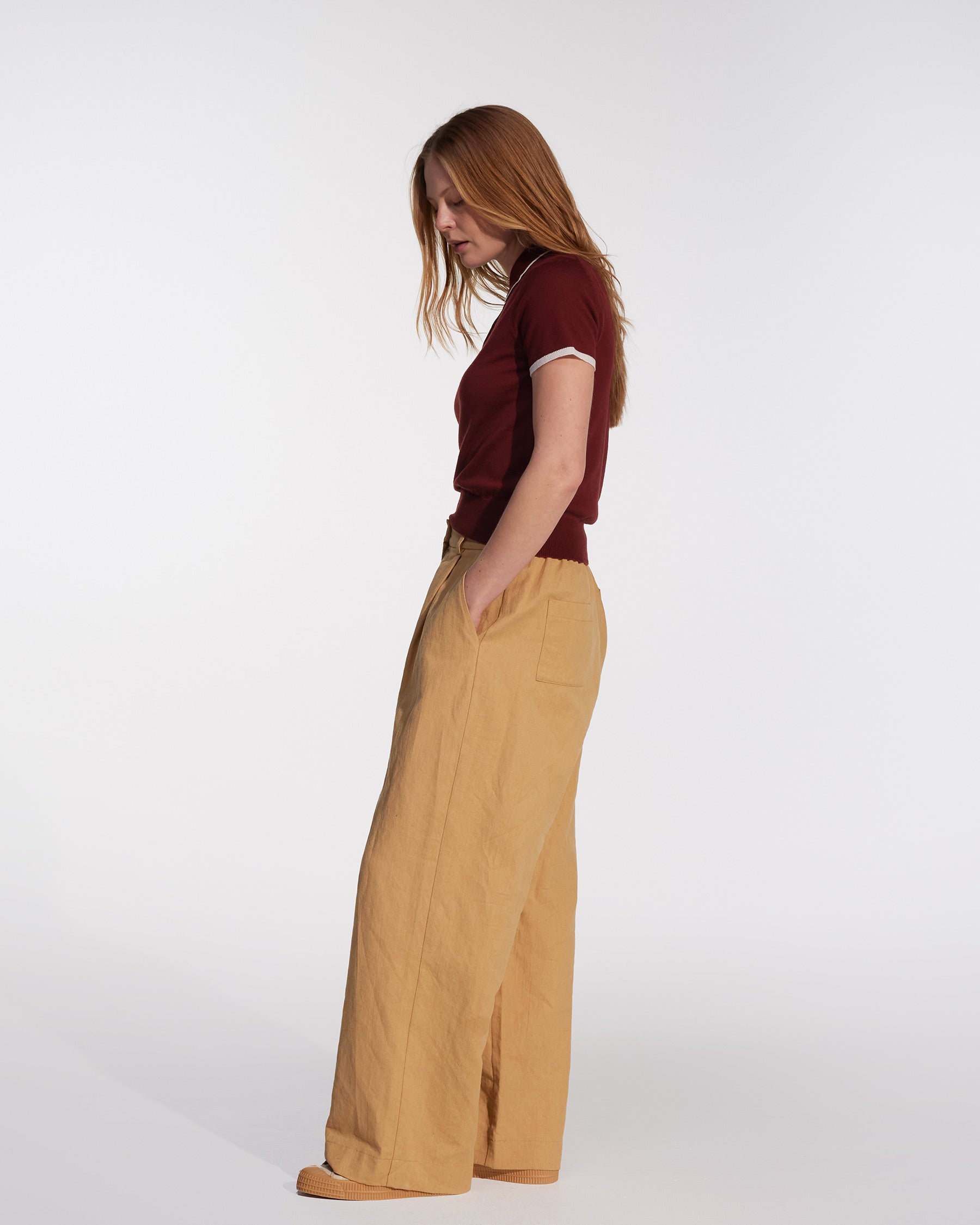 A woman with long reddish hair stands in profile against a plain background, wearing a short-sleeve maroon top and Pyne and Smith Clothiers Cotton Linen Trousers No.16 in Lark Tan, her hands tucked in the pockets.