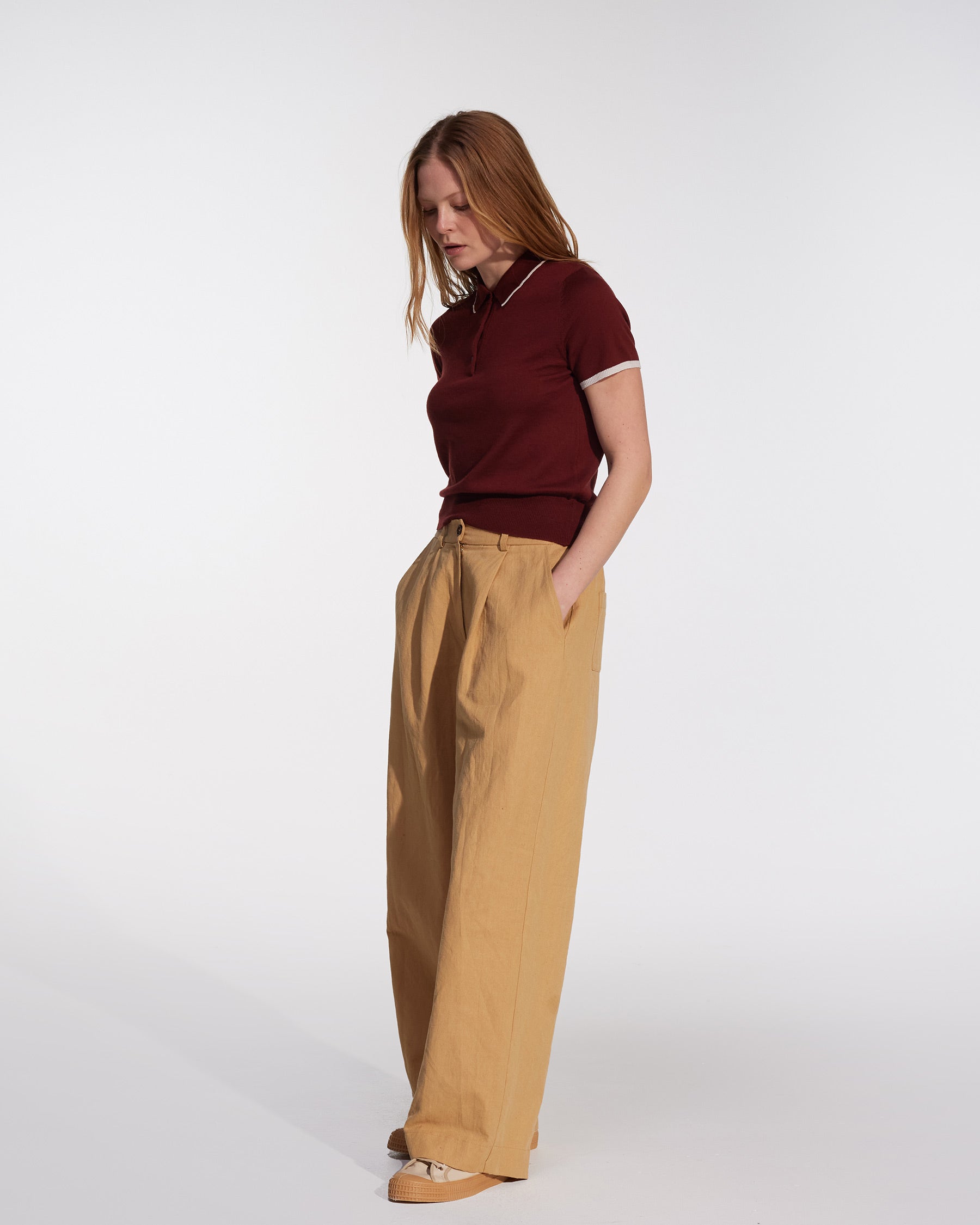 A person with long hair stands against a white background, wearing a maroon polo shirt and Pyne and Smith Clothiers' Cotton Linen Trousers No.16 in Lark Tan, paired with beige shoes. They look down with hands in their pockets.
