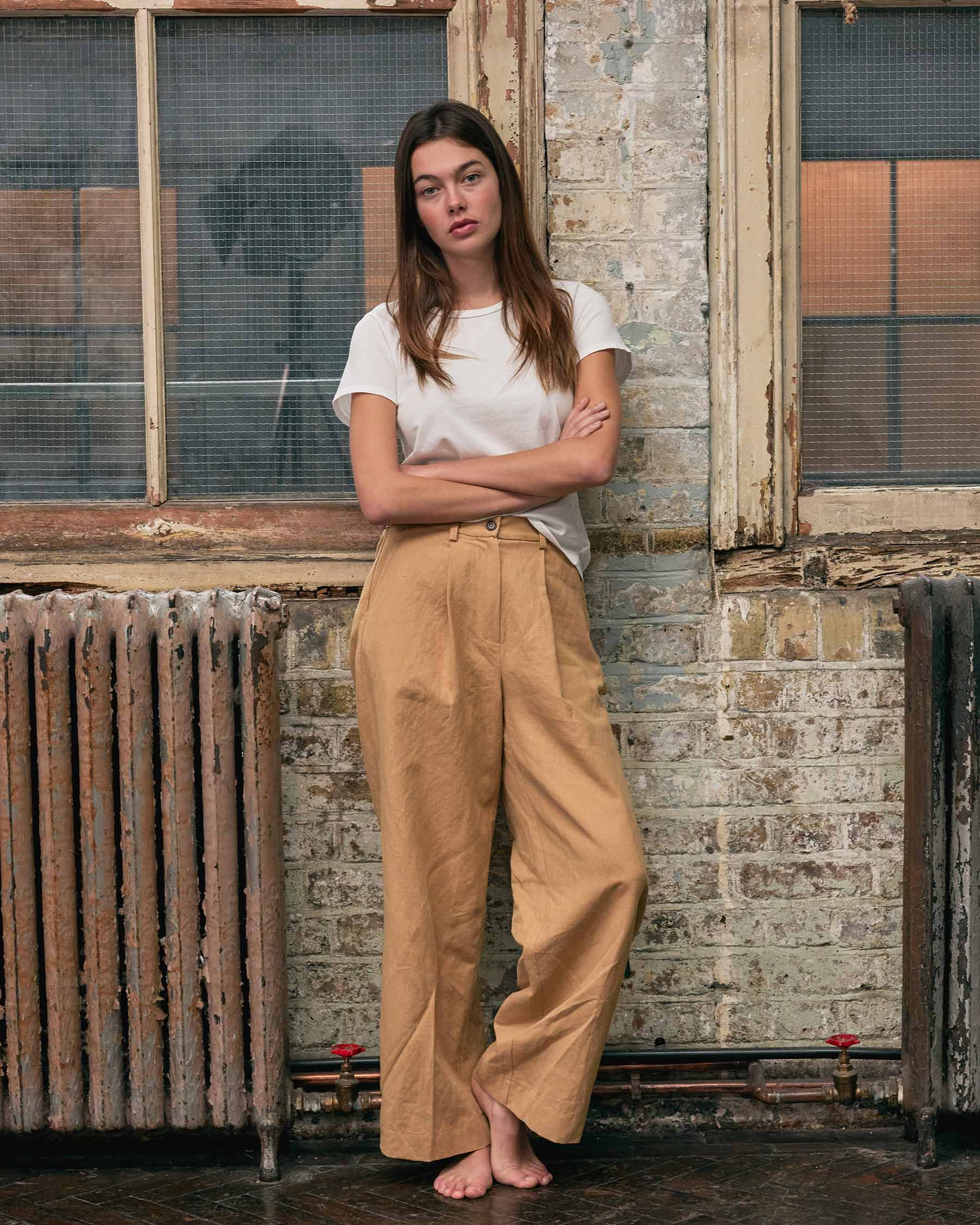 A woman stands indoors, barefoot with arms crossed, wearing a white tee and Pyne and Smith Clothiers’ Cotton Linen Trousers No.16 in Lark Tan. A weathered brick wall, large window, and old radiator form the backdrop.