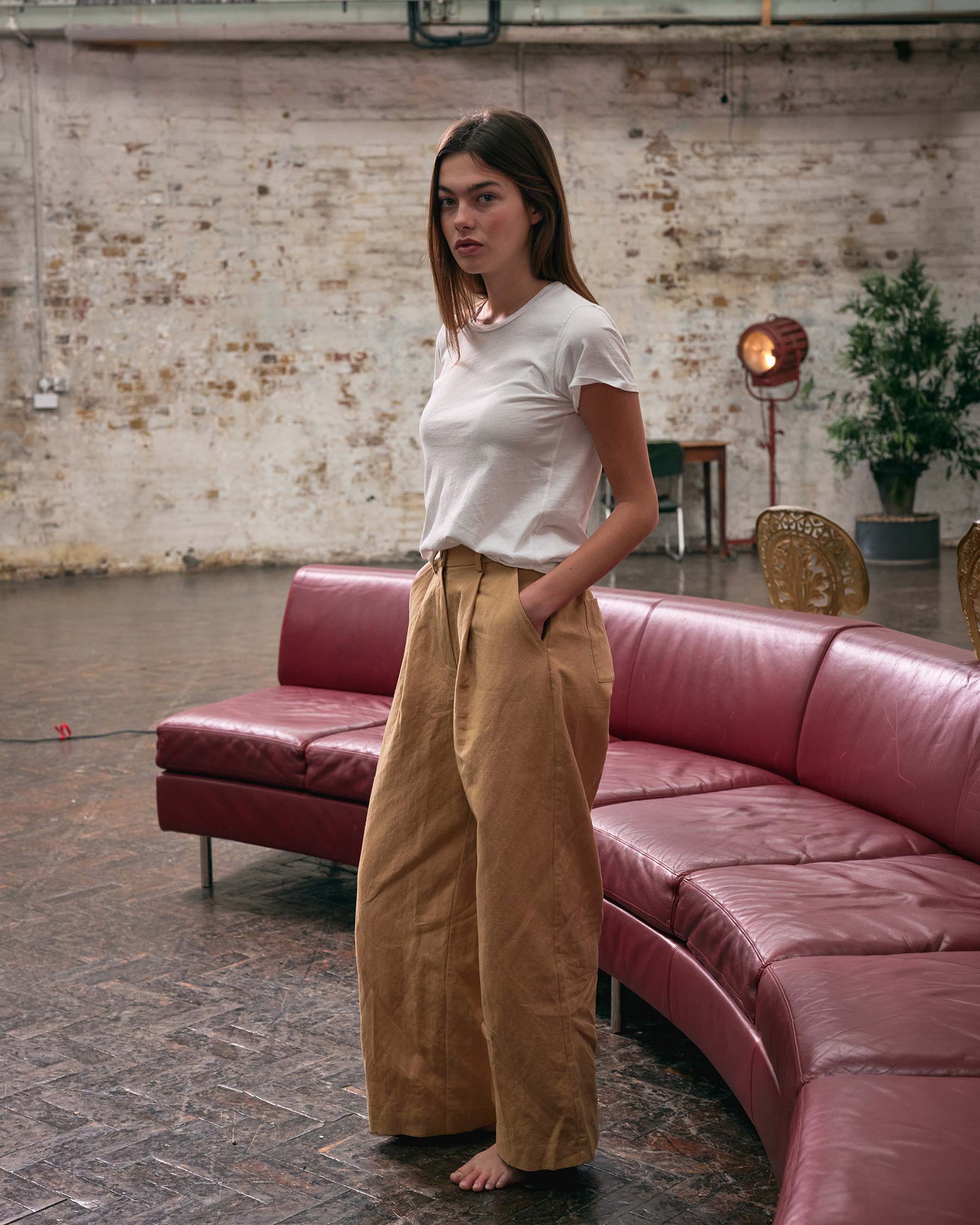A woman with long brown hair stands barefoot on a brick floor, wearing a white tee and Pyne and Smith Clothiers' Cotton Linen Trousers No.16 in Lark Tan beside a curved red couch, with brick wall and vintage lamp in the background.