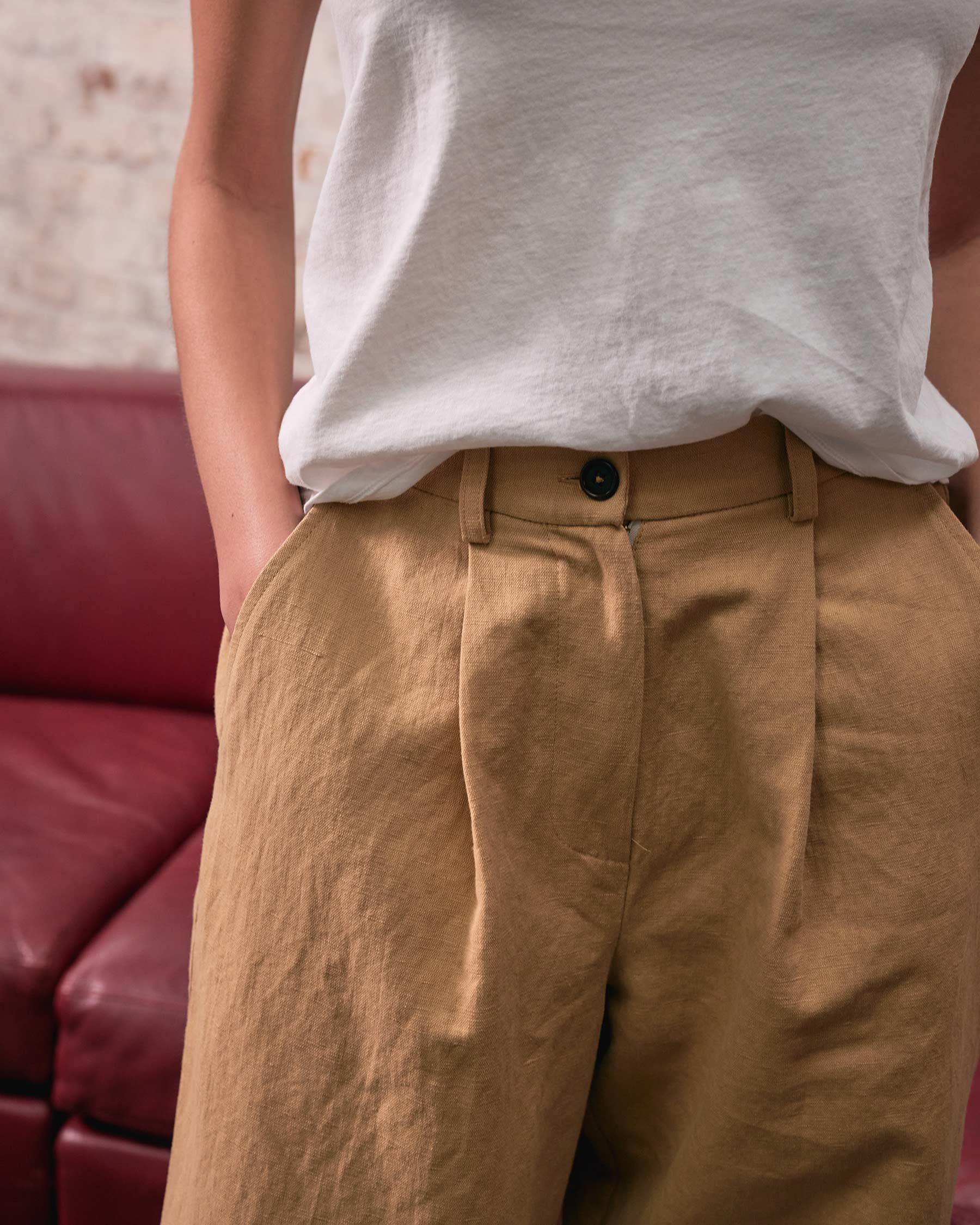 A person wearing a white T-shirt and Pyne and Smith Clothiers Cotton Linen Trousers No.16 in Lark Tan stands with hands in pockets in front of a red couch and a light-colored wall.