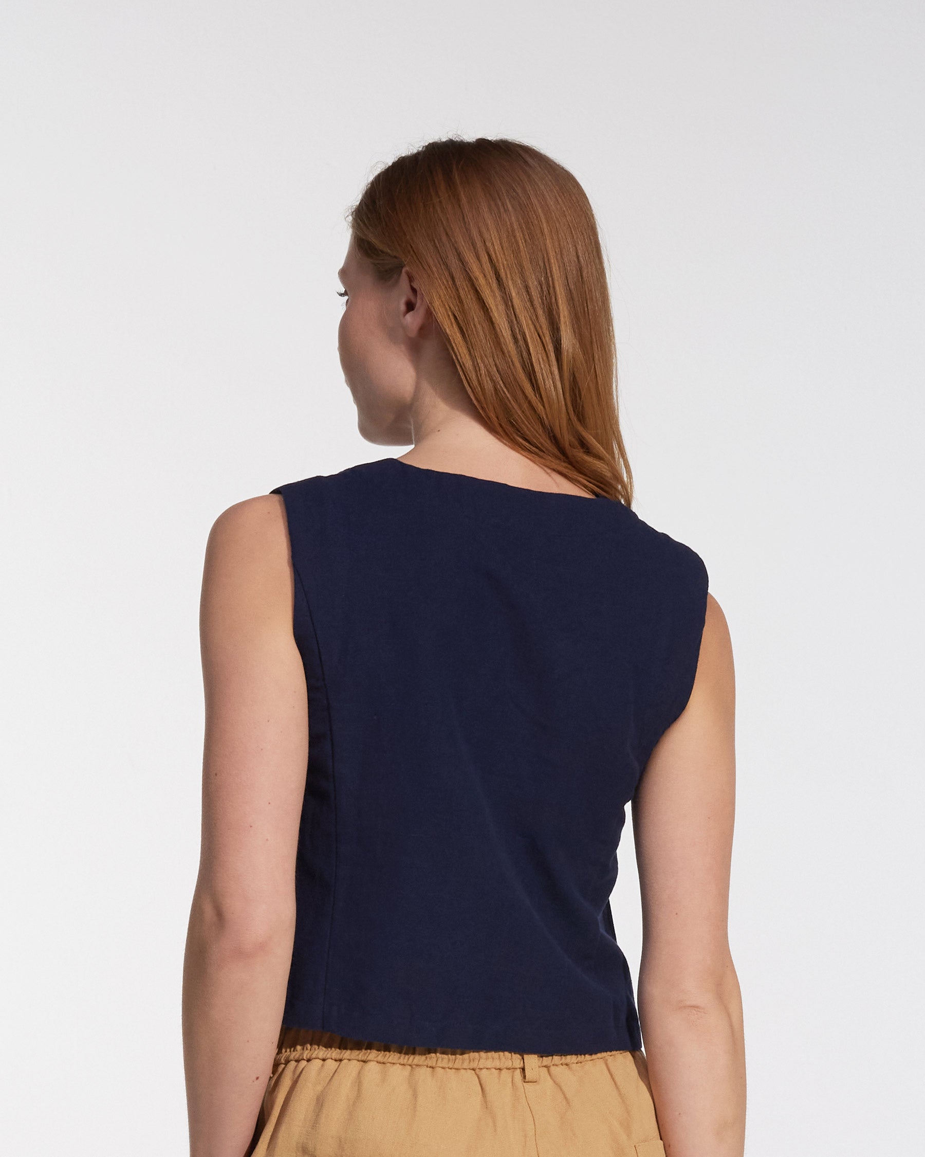 A woman with long, light brown hair is shown from the back wearing the Pyne and Smith Clothiers Button up Vest in Ink Blue Canvas and tan pants against a plain white background.