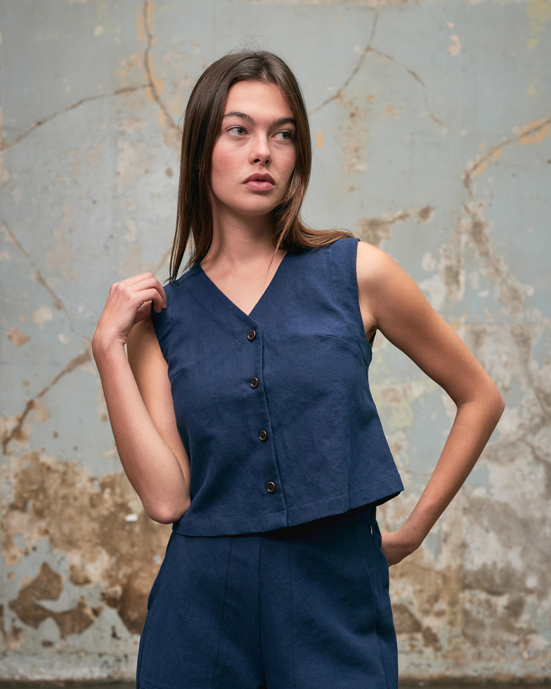 A woman in the Pyne and Smith Clothiers Button up Vest in Ink Blue Canvas stands before a textured wall, looking to the side with one hand touching her collar.