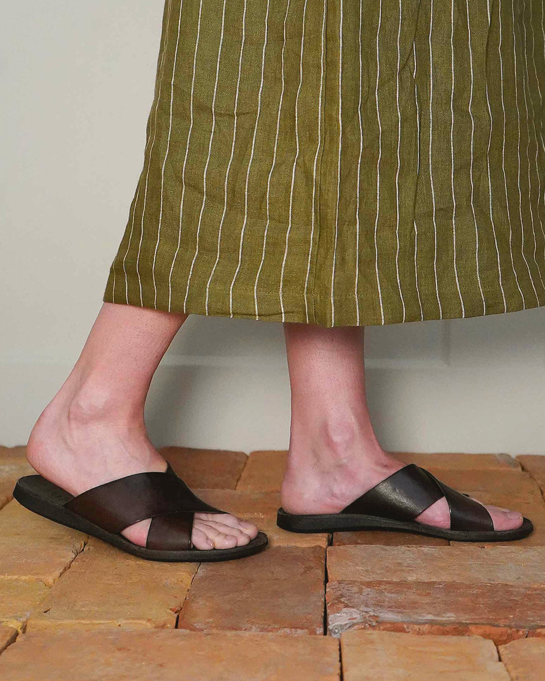 A person in a green striped skirt and Pyne and Smith Clothiers Dark Brown Leather Infinity Sandals stands on reddish-brown brick flooring.
