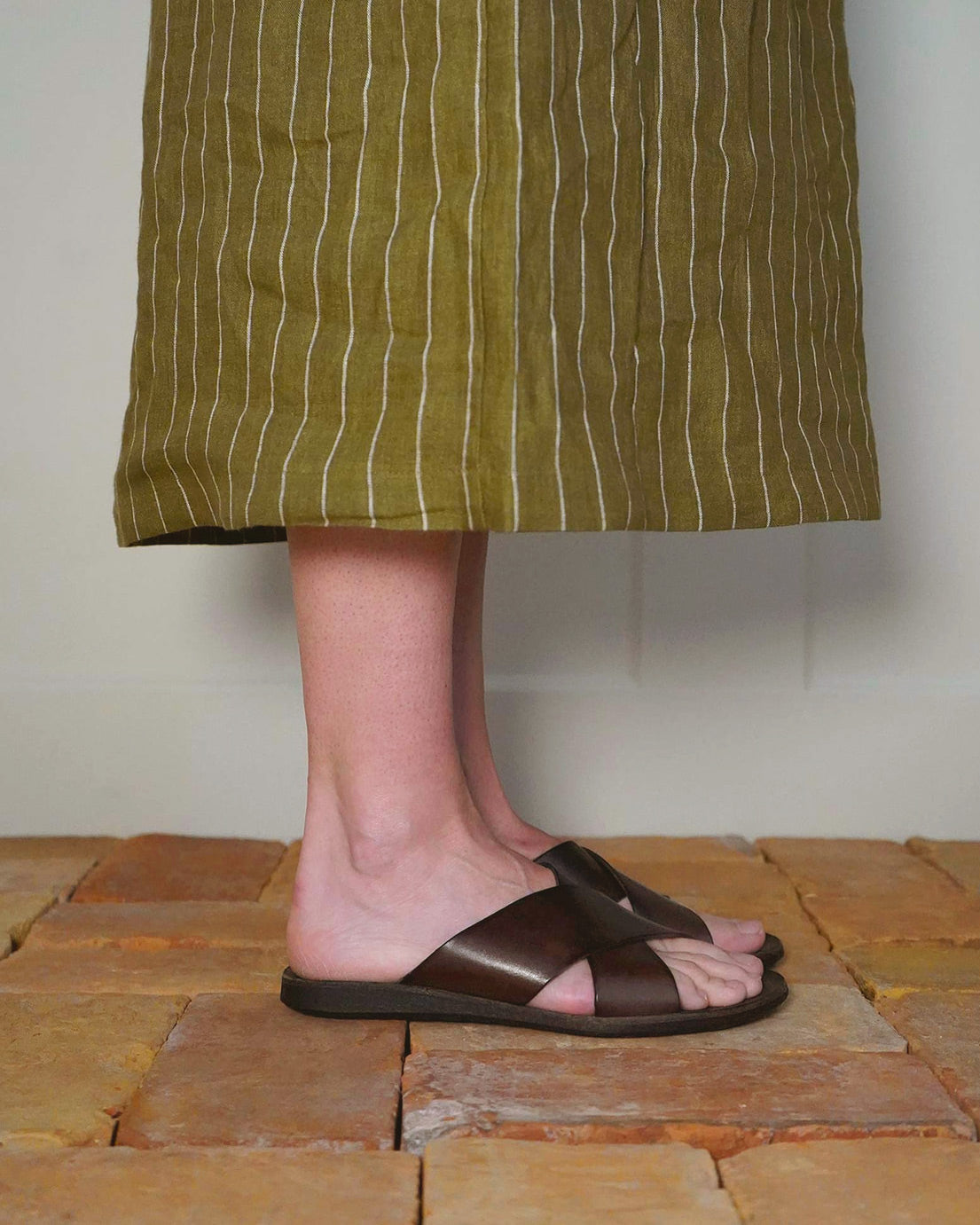 Only the lower legs and feet are visible as a person stands on a tile floor wearing a long, olive-green skirt with white vertical stripes and Pyne and Smith Clothiers’ Dark Brown Leather Infinity Sandal.