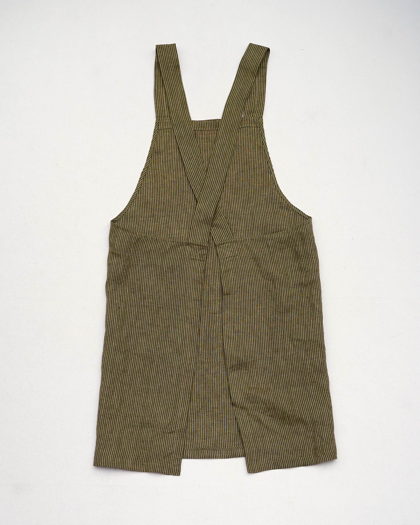 The Cross Back Apron in Olive Stripe Linen by Pyne and Smith Clothiers features wide straps crossed at the back, an open split at the bottom front, and is crafted from ethically made European linen. The apron is shown laid flat on a white surface.