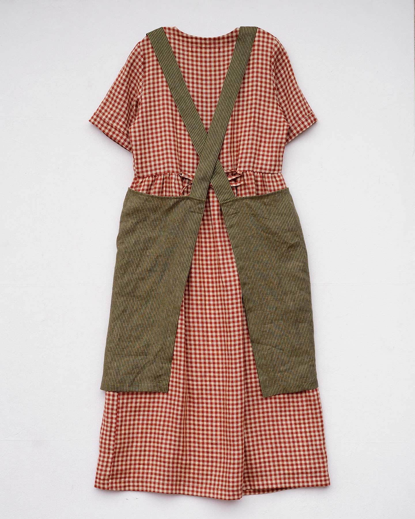 A Cross Back Apron in Olive Stripe Linen by Pyne and Smith Clothiers, featuring wide straps that tie at the waist, is shown layered over a short-sleeved, red and white checkered dress, both laid flat on a white background.