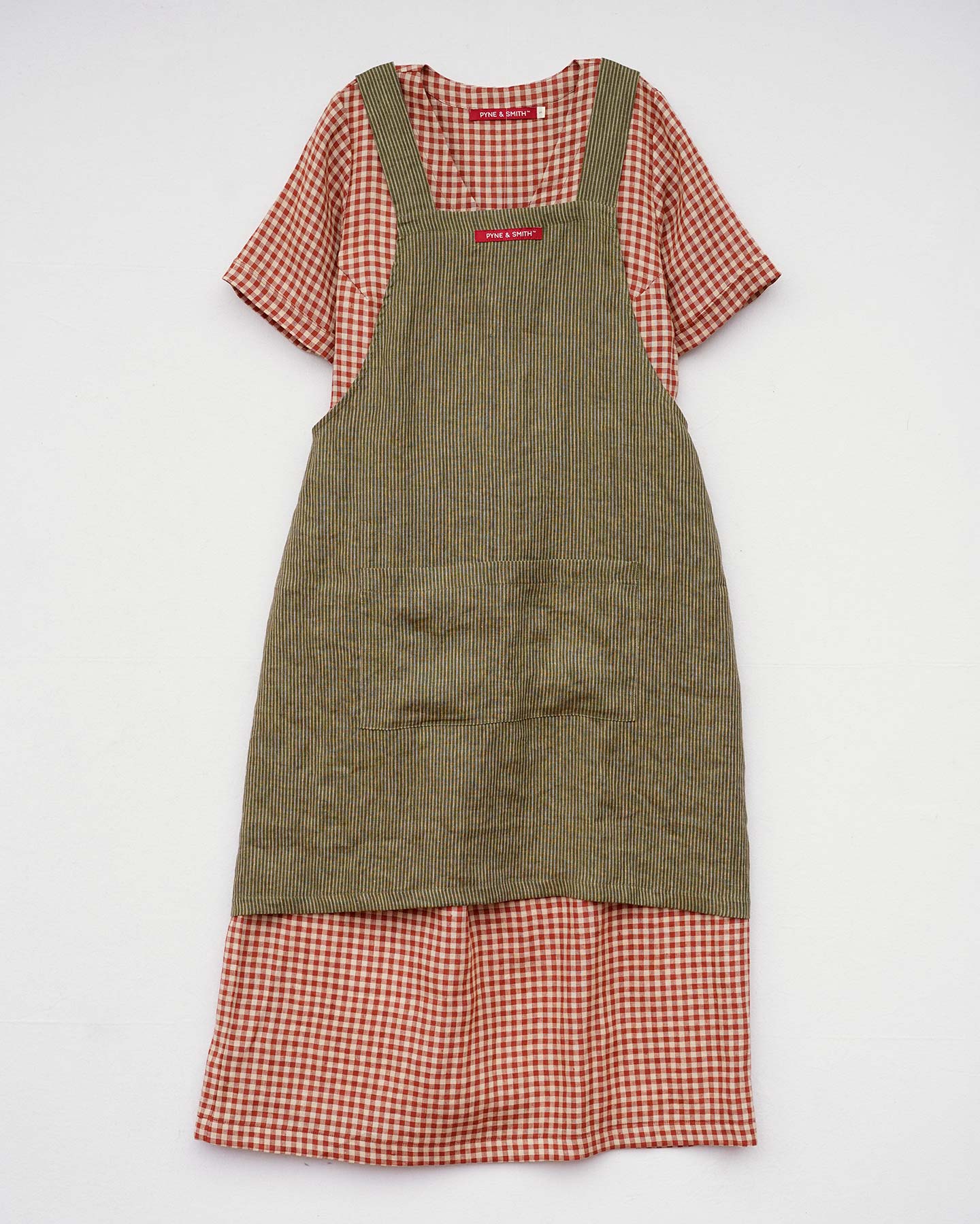 A red and white checkered short-sleeve dress is topped with a Pyne and Smith Clothiers Cross Back Apron in Olive Stripe Linen, both displayed flat on a white background.