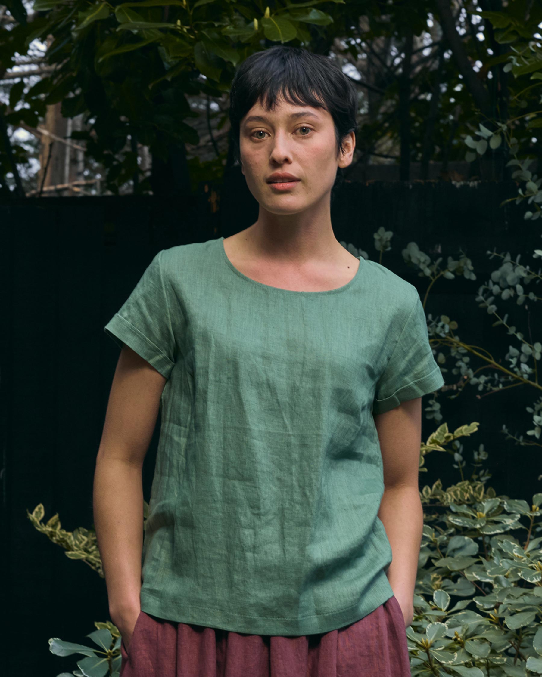 A person with short dark hair stands outdoors among greenery, wearing the Pyne and Smith Clothiers Blouse No.14 Linen T-shirt in Celedon Green and a mauve skirt, creating a serene, sustainable scene.
