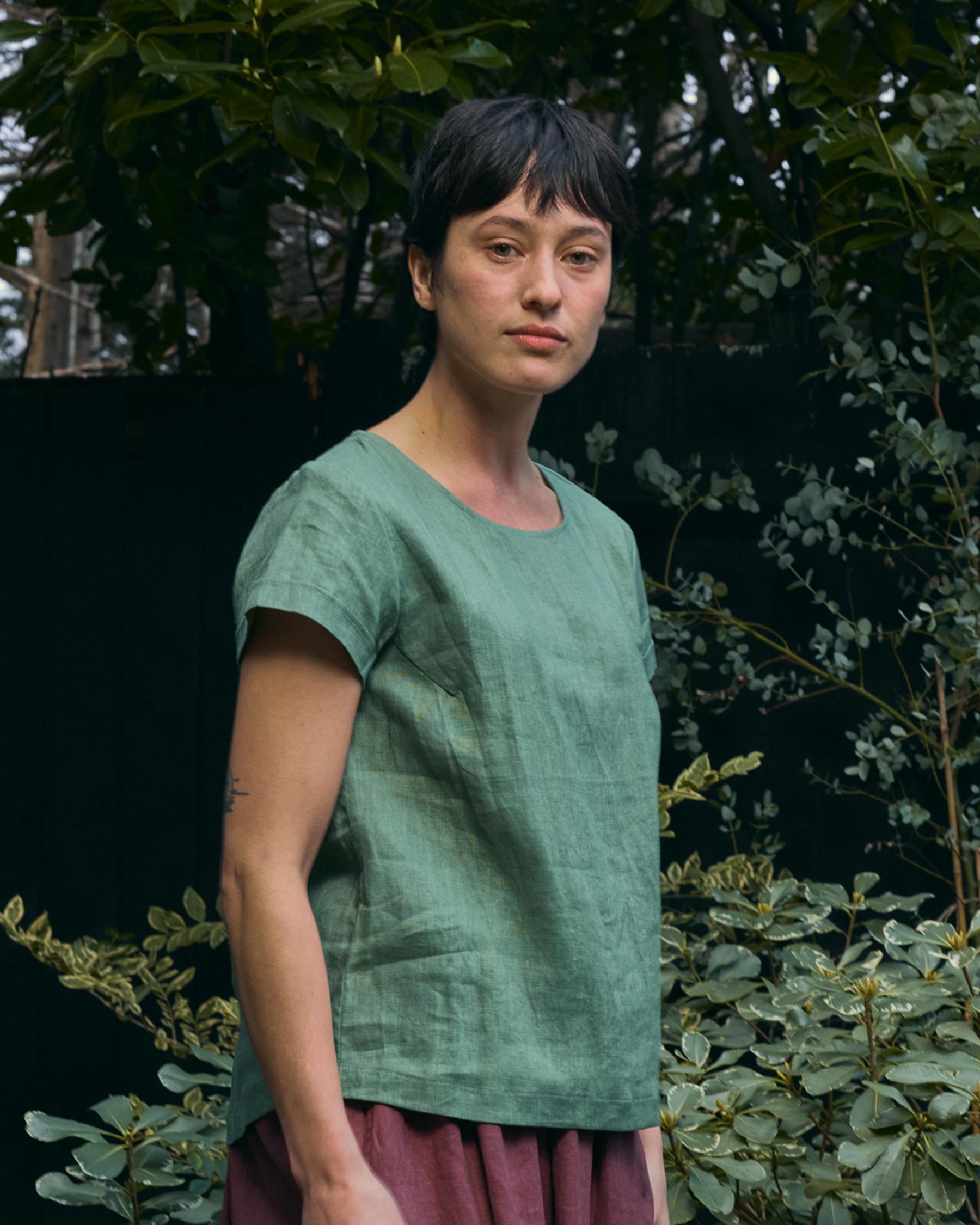 A person with short dark hair wears the Pyne and Smith Clothiers Blouse No.14 Linen T-shirt in Celedon Green and a maroon skirt, standing outdoors before leafy plants, looking calmly at the camera.