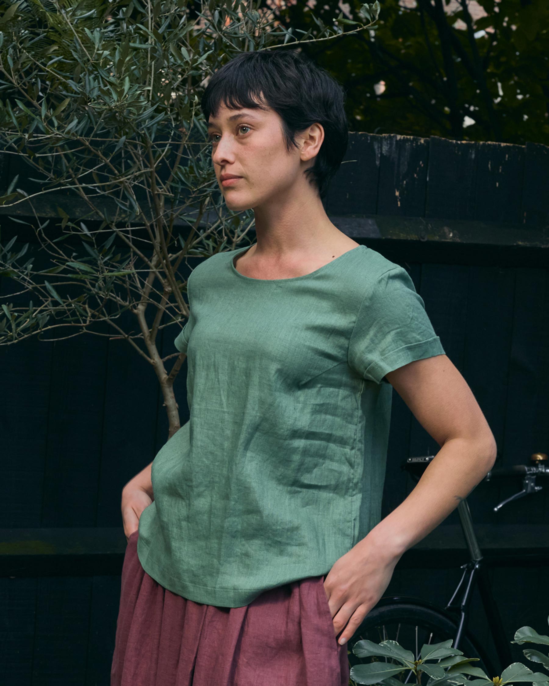 A person with short dark hair wears the Pyne and Smith Clothiers Blouse No.14 Linen T-shirt in Celedon Green and a mauve skirt, standing outdoors by plants and a dark wooden fence, with a bicycle partly visible in the background.