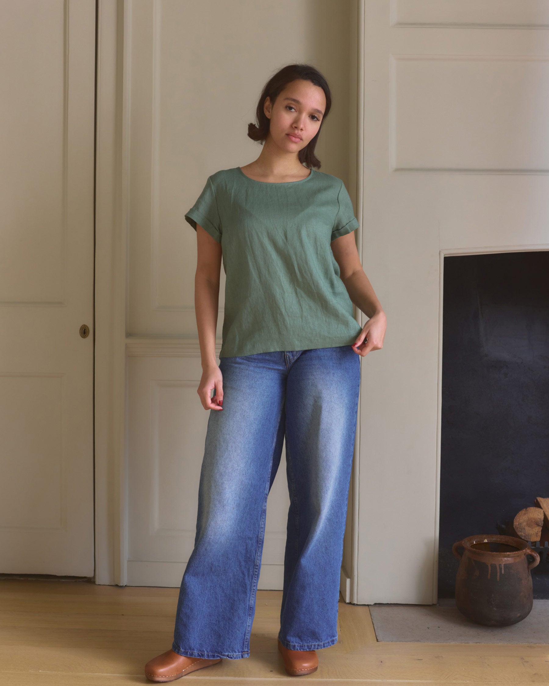 A woman stands indoors wearing the Pyne and Smith Clothiers Blouse No.14 Linen T-shirt in Celedon Green, wide-leg blue jeans, and brown shoes, posing casually by a white wall and fireplace with pottery on the floor.