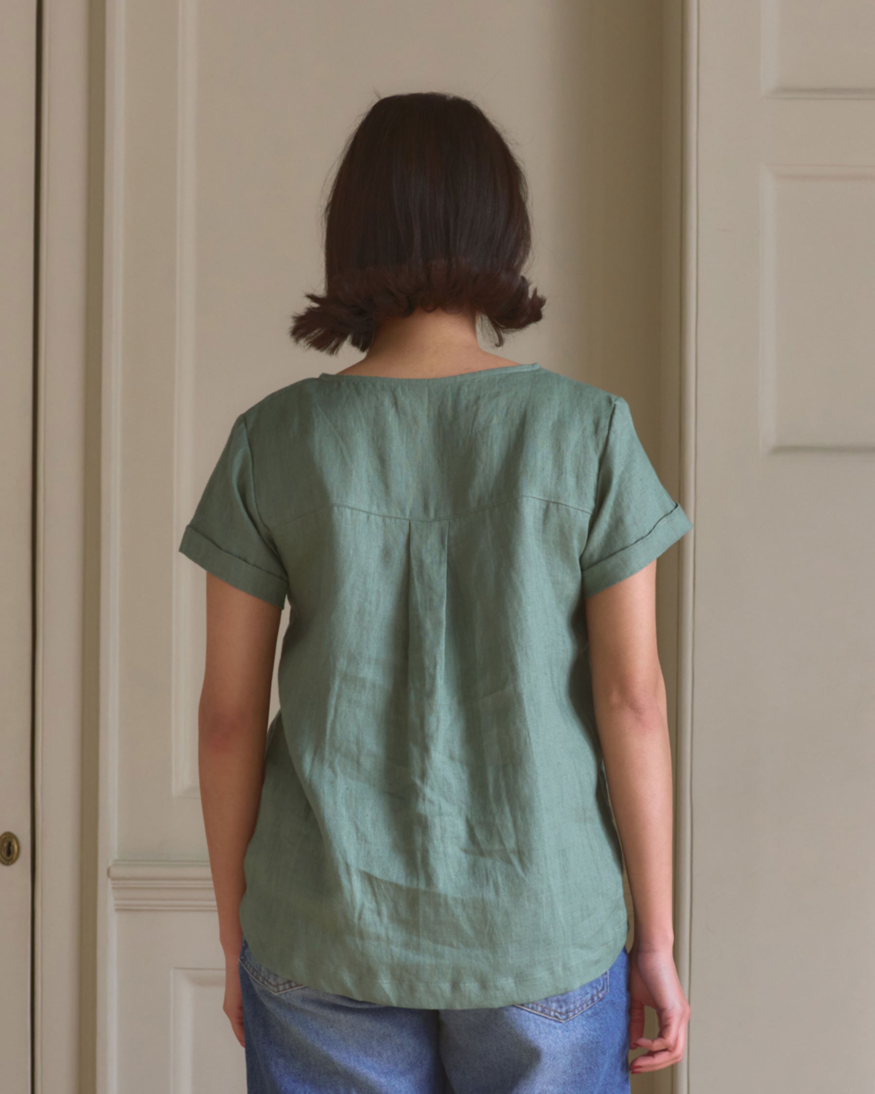 A person with short brown hair, wearing the Pyne and Smith Clothiers Blouse No.14 Linen T-shirt in Celedon Green and blue jeans, stands with their back to the camera in front of a light-colored wall.