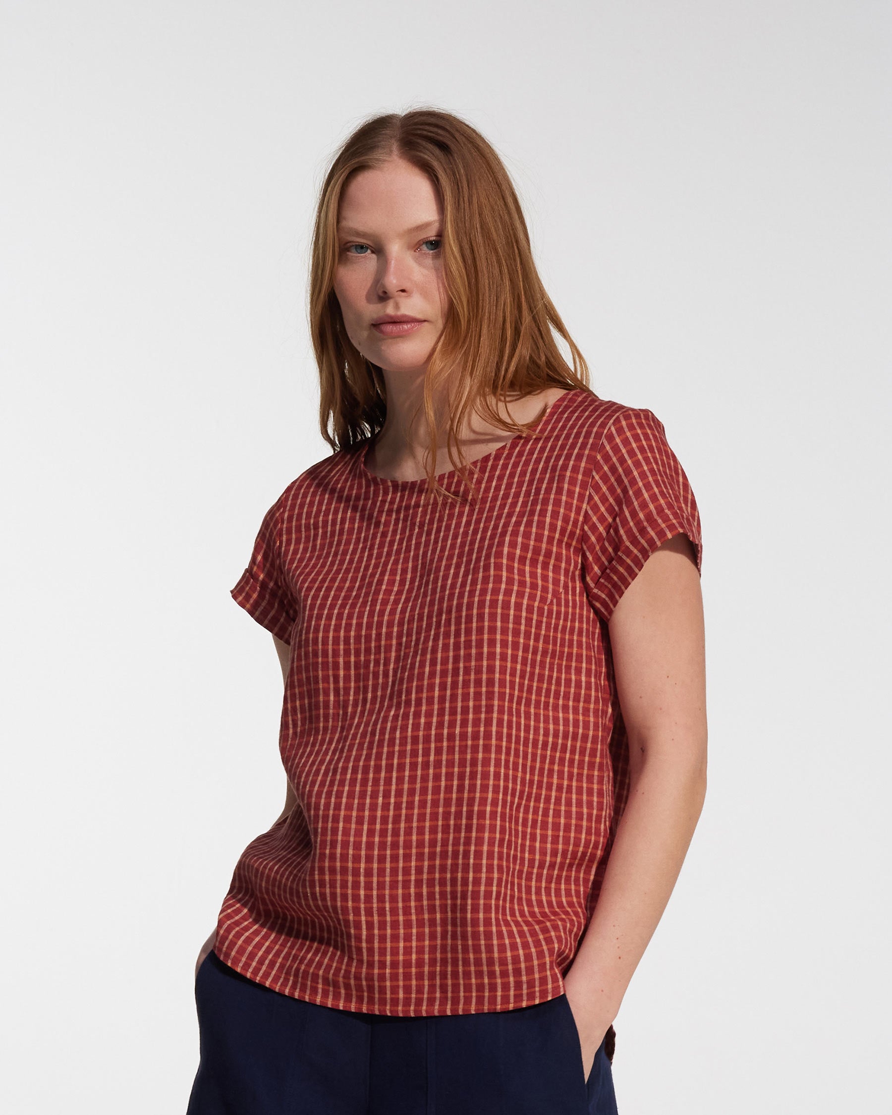 A woman with long, straight, light brown hair wears the Pyne and Smith Clothiers Blouse No.14 Linen T-shirt in Pomodoro Plaid and dark pants, standing with hands in her pockets against a plain white background.