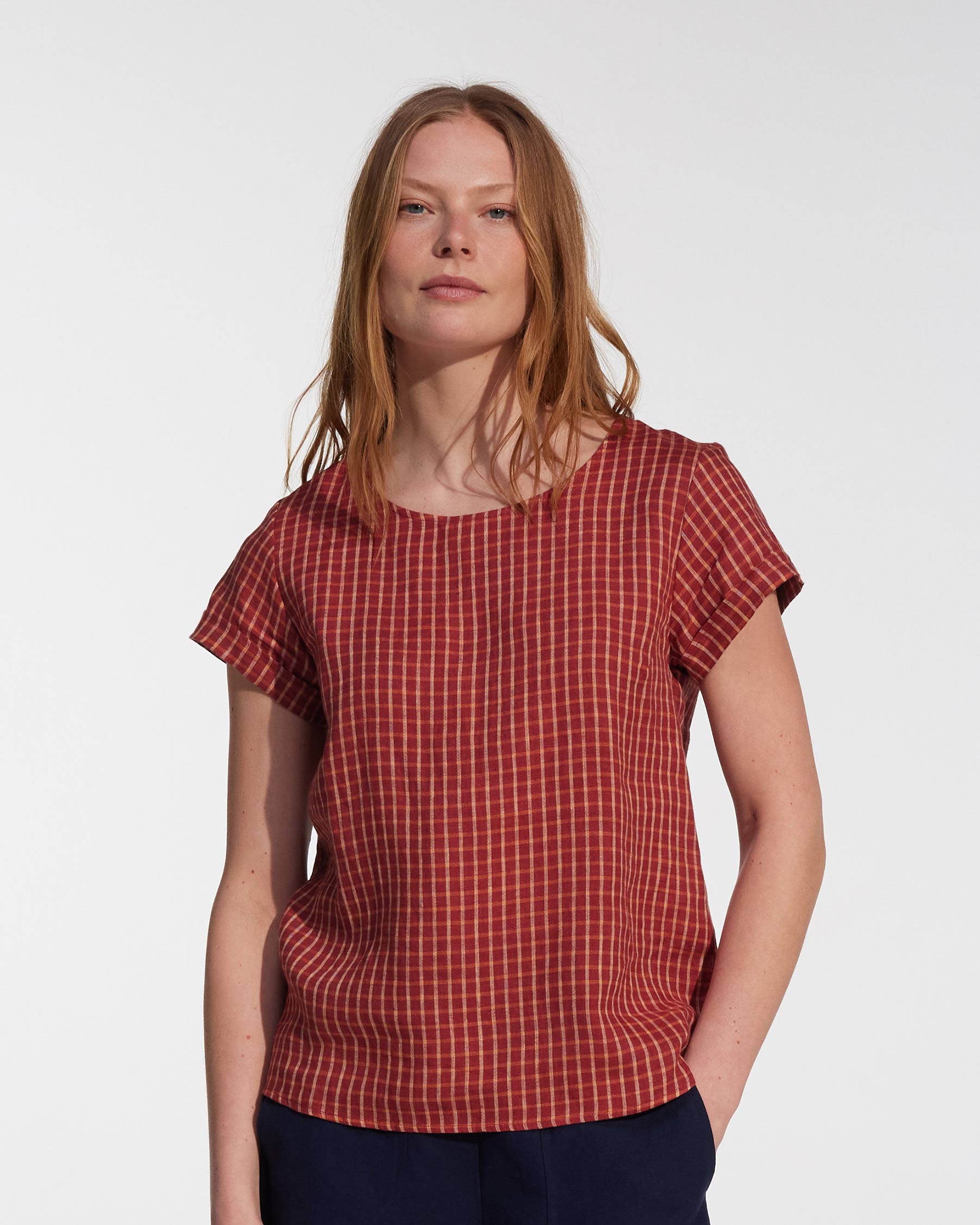 A woman with long, straight, light brown hair wears the Pyne and Smith Clothiers Blouse No.14 Linen T-shirt in Pomodoro Plaid with dark pants, standing against a plain white background.