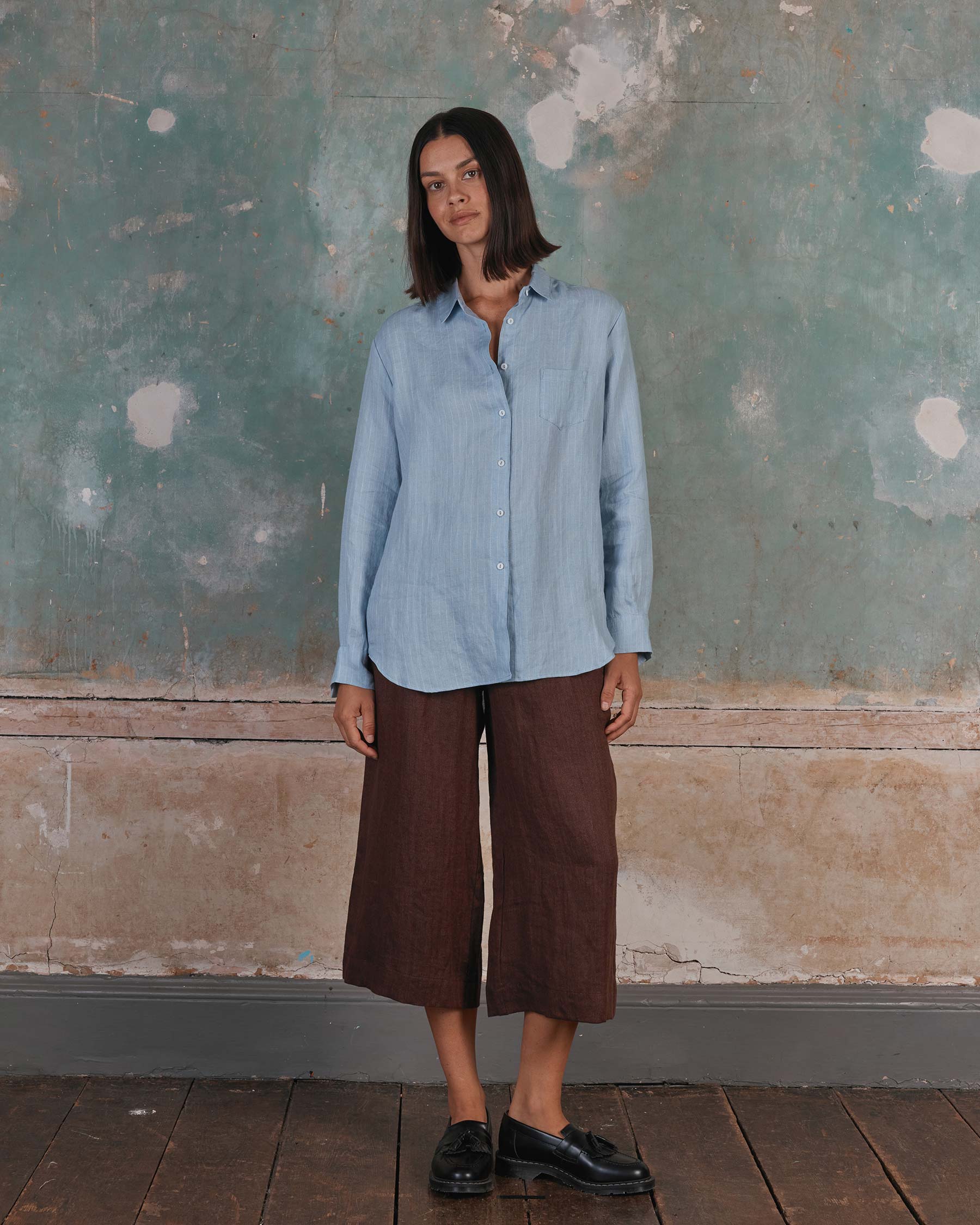 A woman stands against a textured wall, wearing Pyne and Smith Clothiers’ Shirt No.21 Button Up, a linen blouse in pale blue pinstripe, with loose brown culottes and black shoes. Her straight shoulder-length hair frames her neutral expression.