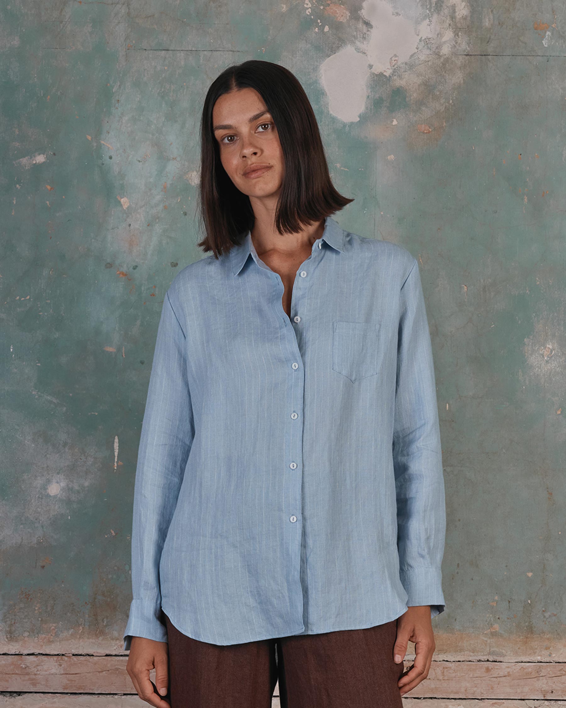 A woman with straight brown hair wears the Pyne and Smith Clothiers Shirt No.21 Button up, a pale blue pinstripe linen blouse, and brown pants as she stands calmly before a weathered green wall with peeling paint.
