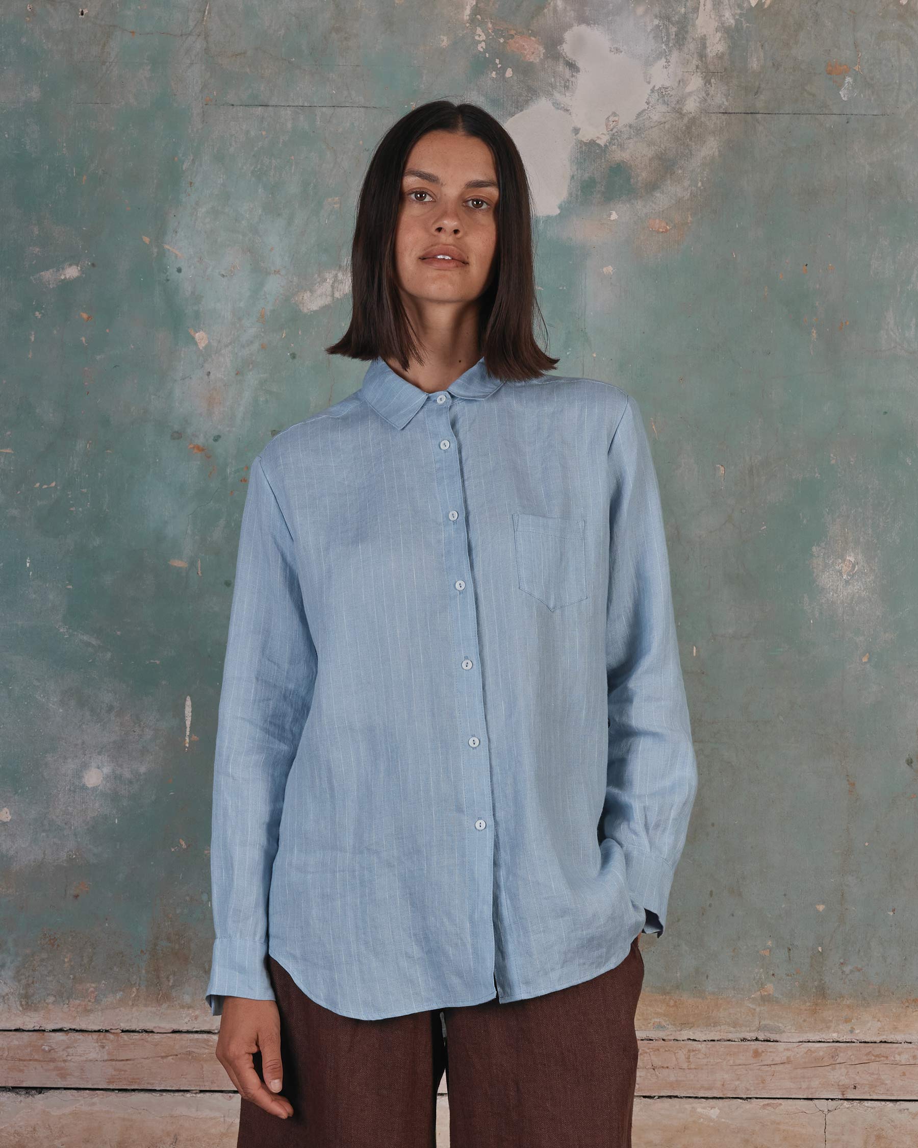 A woman with straight brown hair stands against a faded green and white wall, wearing the Pyne and Smith Clothiers Shirt No.21 Button up—a pale blue pinstripe linen blouse—paired with brown pants, one hand in her pocket as she looks calmly at the camera.