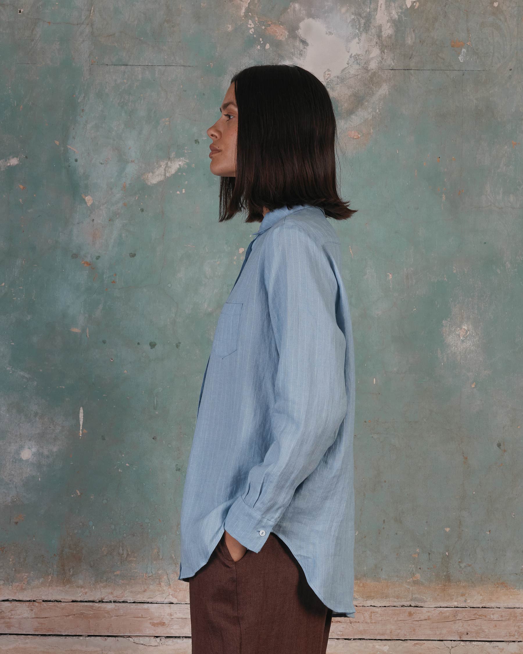 A person with shoulder-length dark hair, wearing the Pyne and Smith Clothiers Shirt No.21 Button up, Linen Blouse in Pale Blue Pinstripe and brown pants, stands in profile against a textured teal wall with peeling paint.