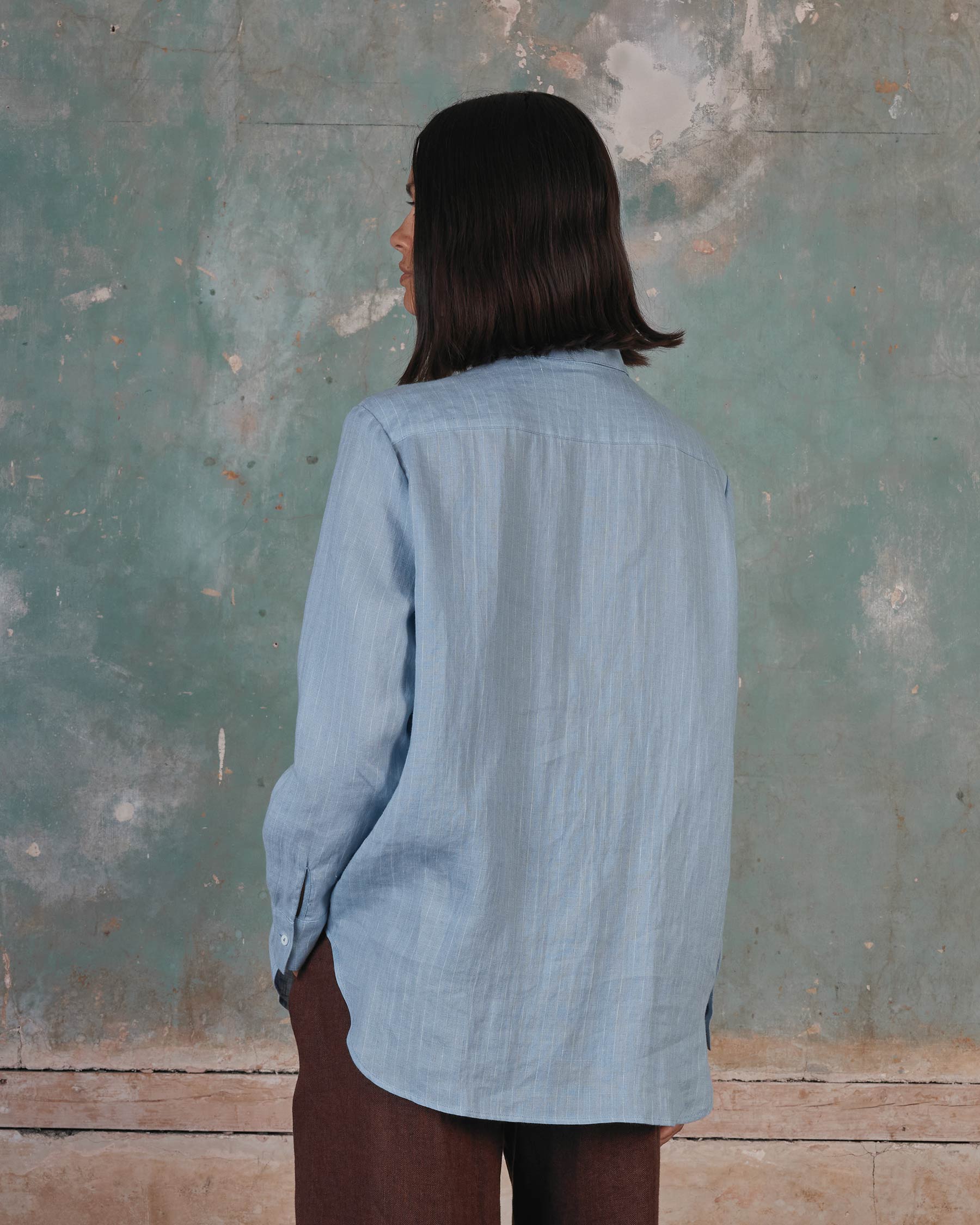 A person with shoulder-length dark hair wears the Pyne and Smith Clothiers Shirt No.21 Button up, a linen blouse in pale blue pinstripe, standing against a textured, weathered blue-green wall with their hands in their pockets.