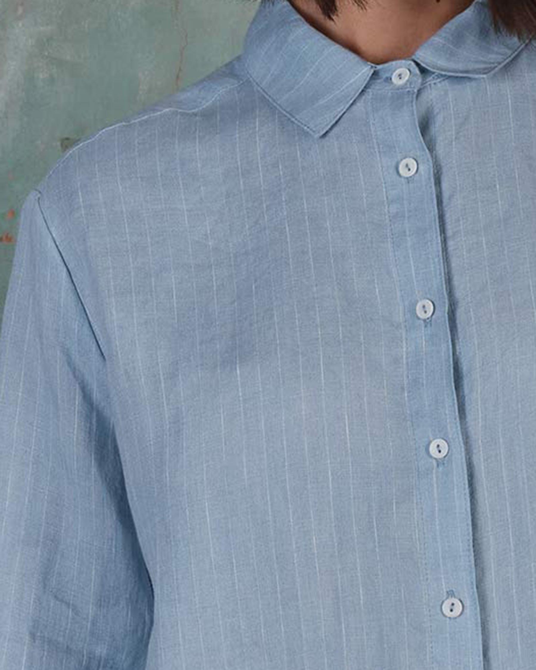 Close-up of a person wearing Pyne and Smith Clothiers Shirt No.21 Button up, a premium European linen blouse in pale blue pinstripe with a collar and white buttons. Thin vertical stripes stand out against the textured, muted green background.