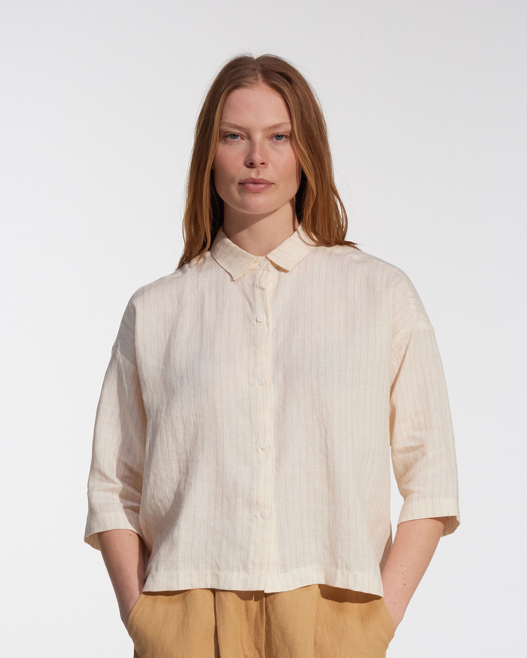 A woman with straight, light brown hair wears the Pyne and Smith Clothiers Boxy Cropped Linen Blouse No.22 in Cream Pinstripe and tan pants, standing against a plain white background with a neutral expression.