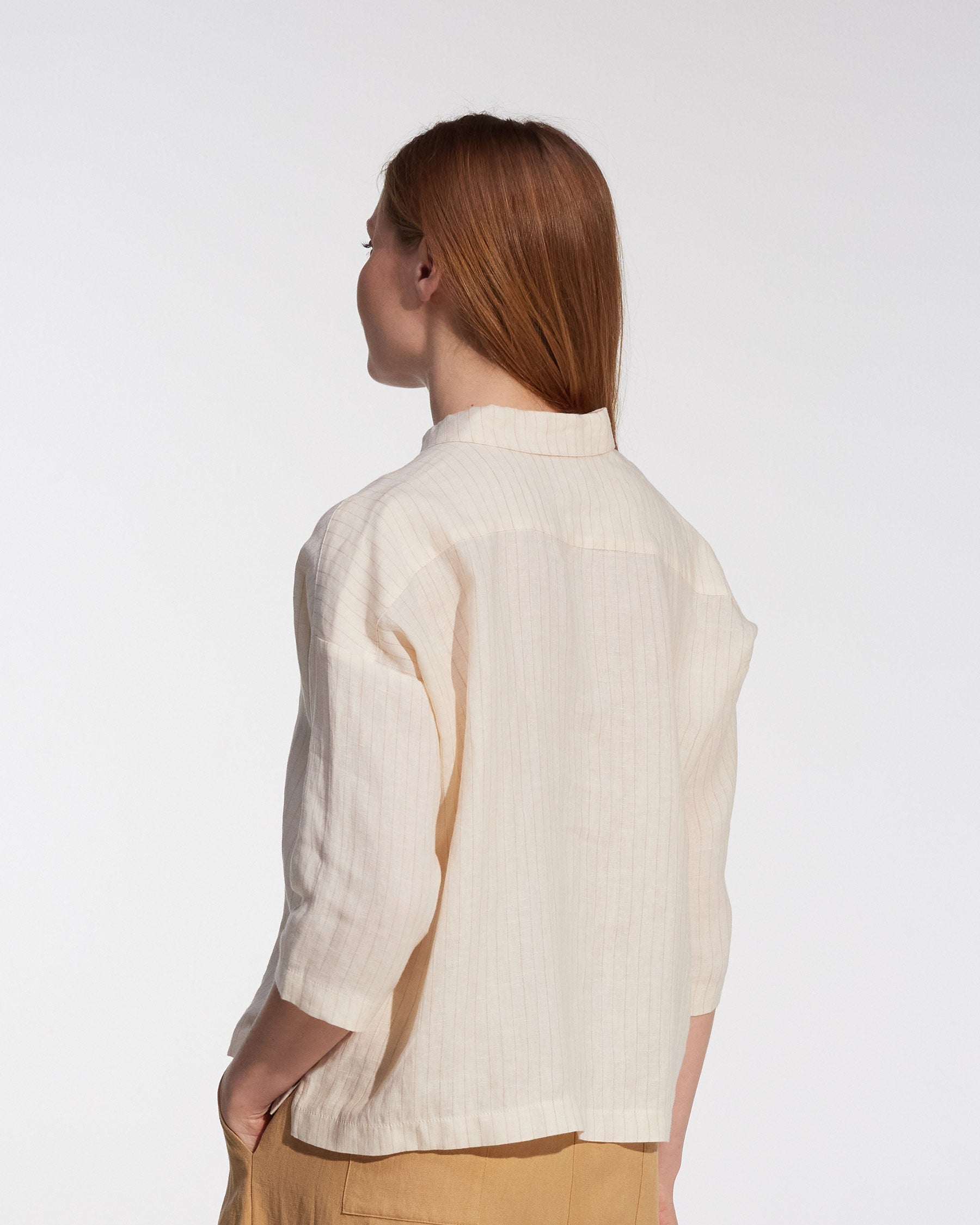 A person with straight, long reddish-brown hair stands sideways, wearing the Pyne and Smith Clothiers Boxy Cropped Linen Blouse No.22 in Cream Pinstripe and light brown pants against a plain white background.
