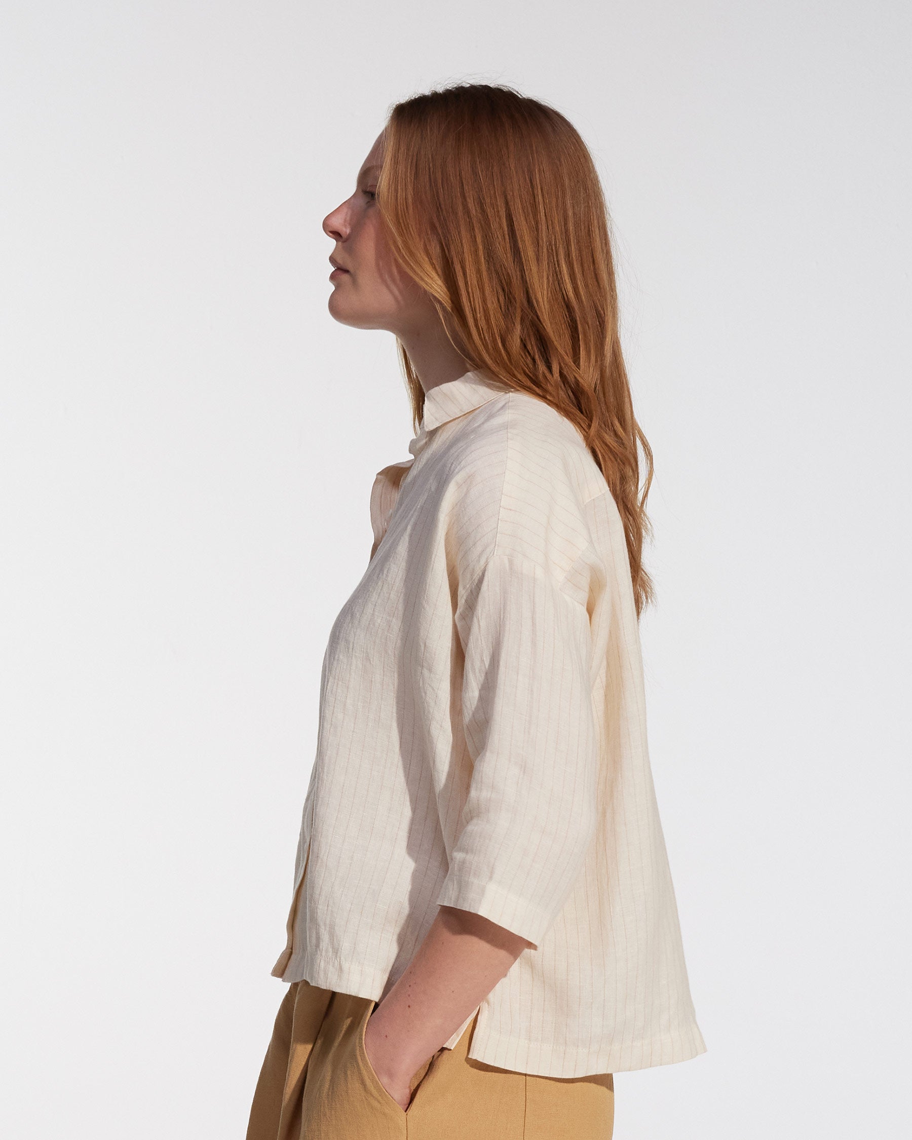 A woman with long, straight, reddish-brown hair stands in profile against a light background, wearing Pyne and Smith Clothiers’ Boxy Cropped Linen Blouse No.22 in Cream Pinstripe and light brown pants, hands casually in her pockets.