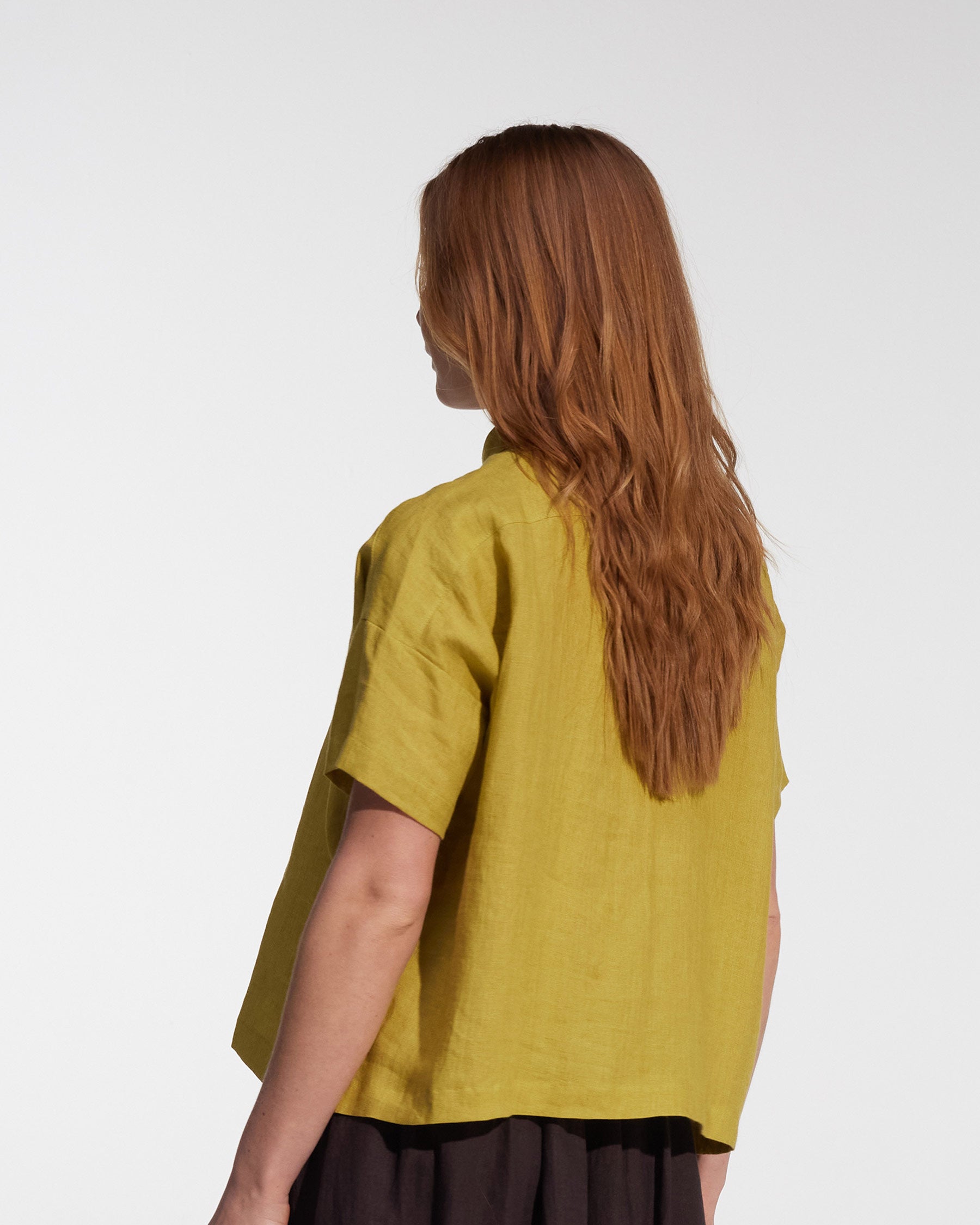 A person with long, wavy, light brown hair is shown from behind in the Cropped Linen Blouse No.24 in Apple by Pyne and Smith Clothiers, paired with a dark skirt, standing against a plain white background.