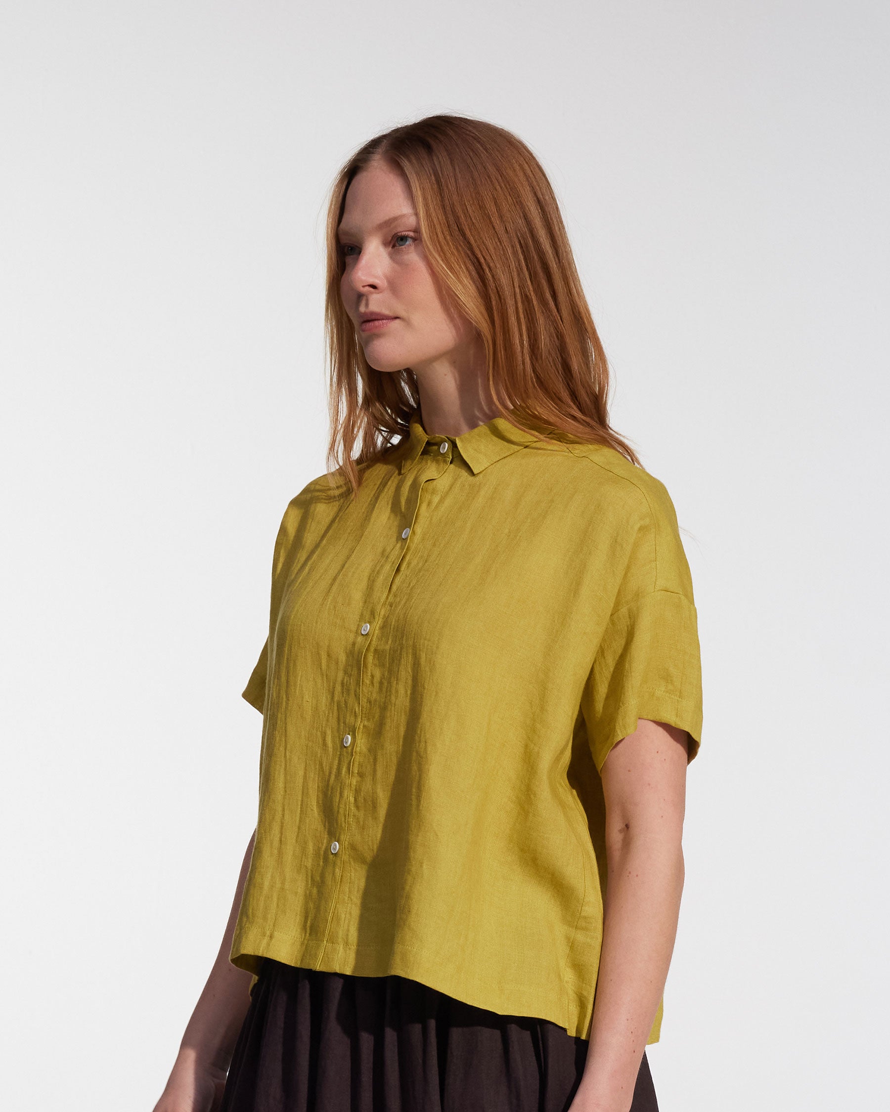 A woman with long, straight, light brown hair wears the Pyne and Smith Clothiers Cropped Linen Blouse No.24 in Apple and a dark skirt, standing against a plain white background.