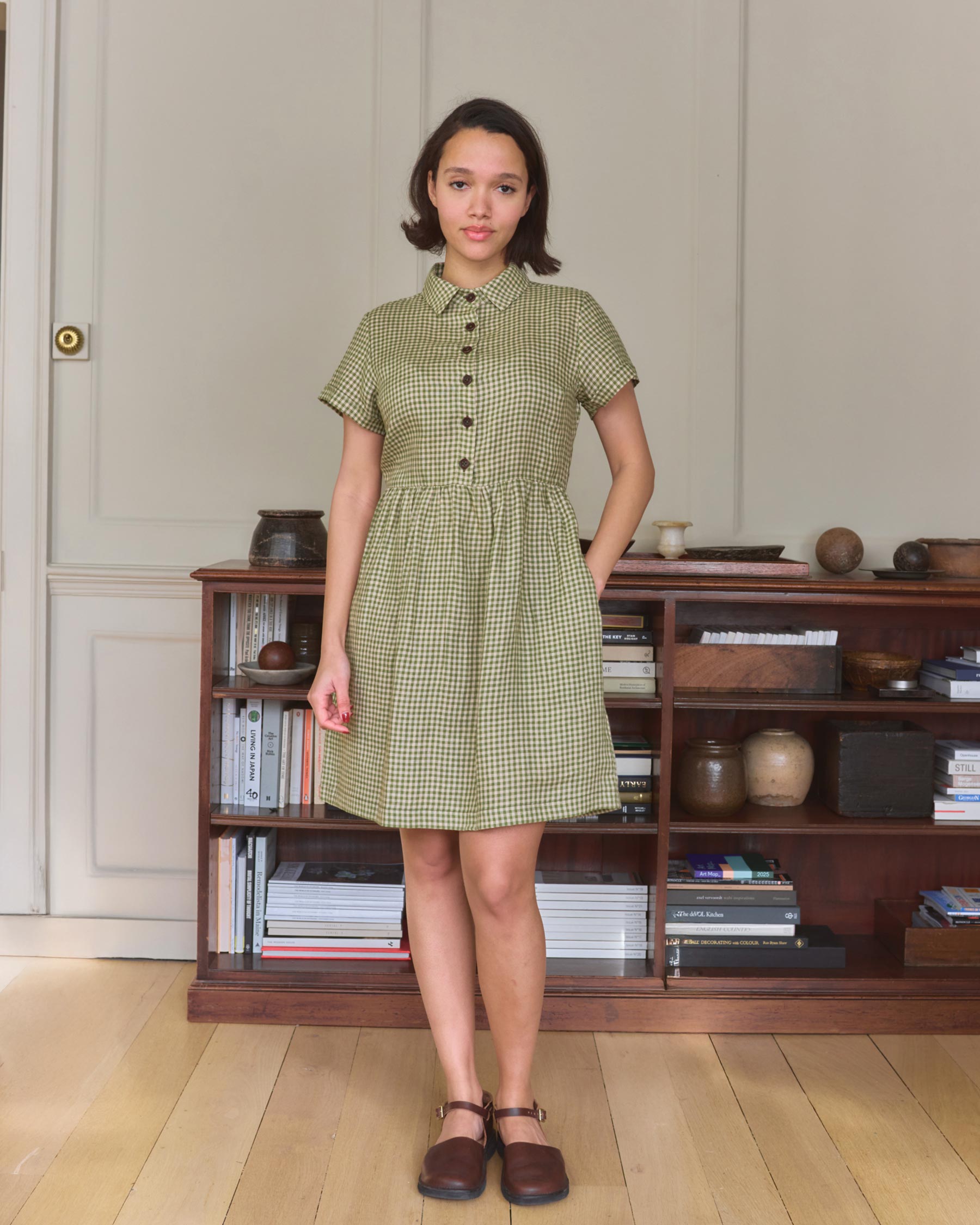 A young woman with short brown hair wears the No.23 Collared, Knee Length Linen Dress in Wimbledon by Pyne and Smith Clothiers, standing indoors on a wooden floor near a bookshelf filled with books and decor, paired with brown shoes.