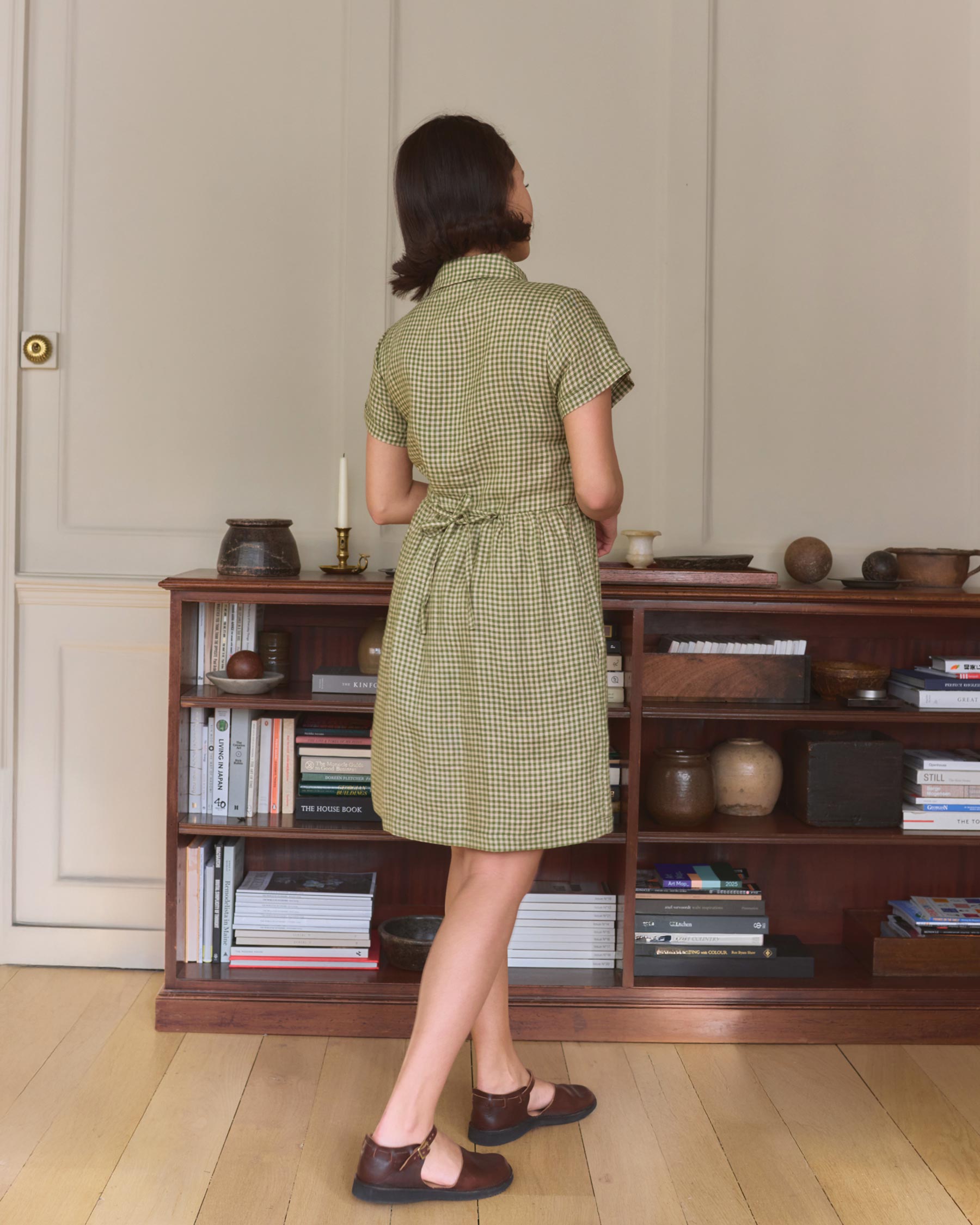 A woman stands barefoot on a wooden floor in a softly lit room, wearing the No.23 Collared, Knee Length Linen Dress in Wimbledon by Pyne and Smith Clothiers while facing a bookshelf filled with books and decorative objects.