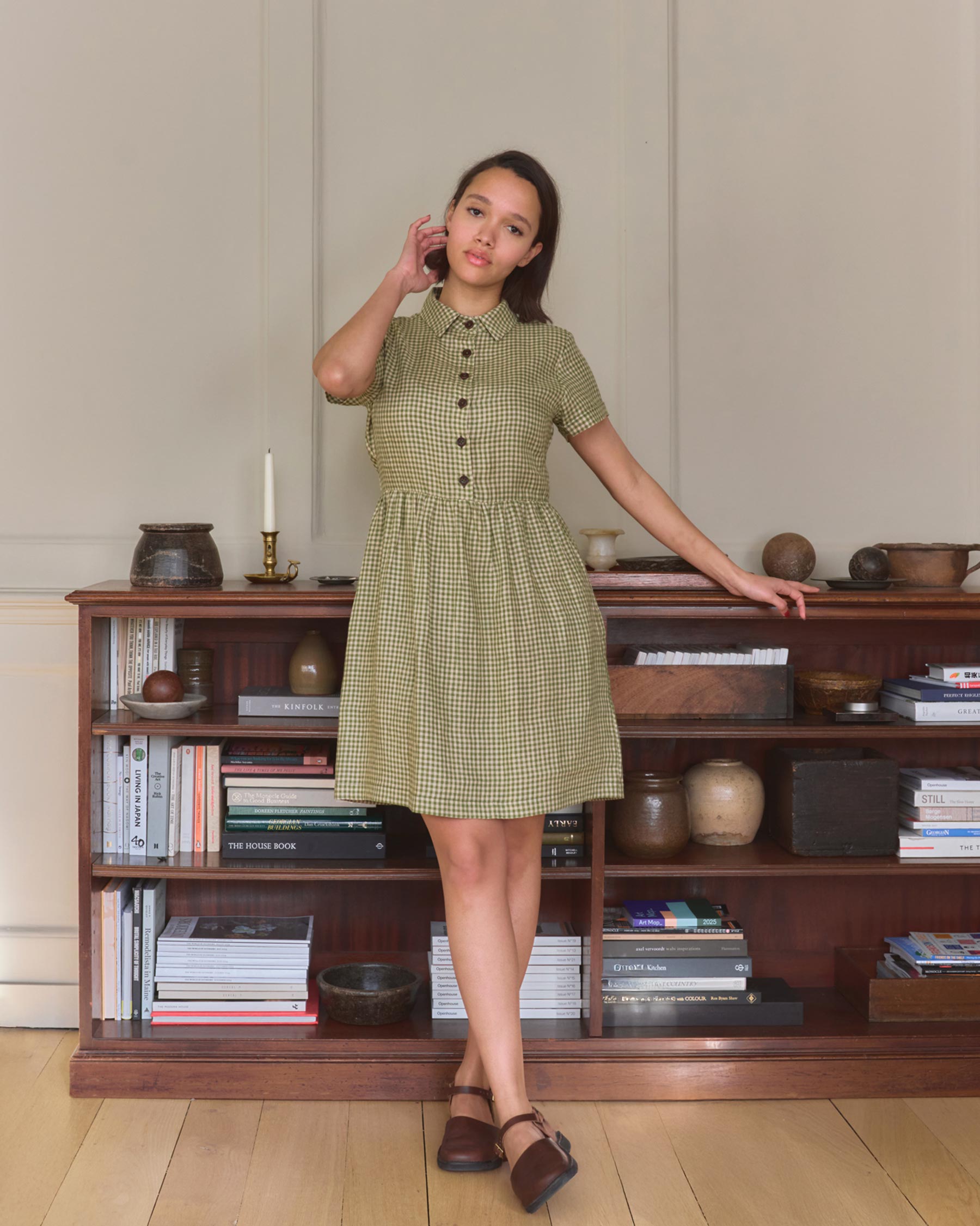 A young woman wears the No.23 Collared, Knee Length Linen Dress in Wimbledon by Pyne and Smith Clothiers, featuring an adjustable waist tie. She stands indoors on a wooden floor beside a bookshelf with books, pottery, and decor.