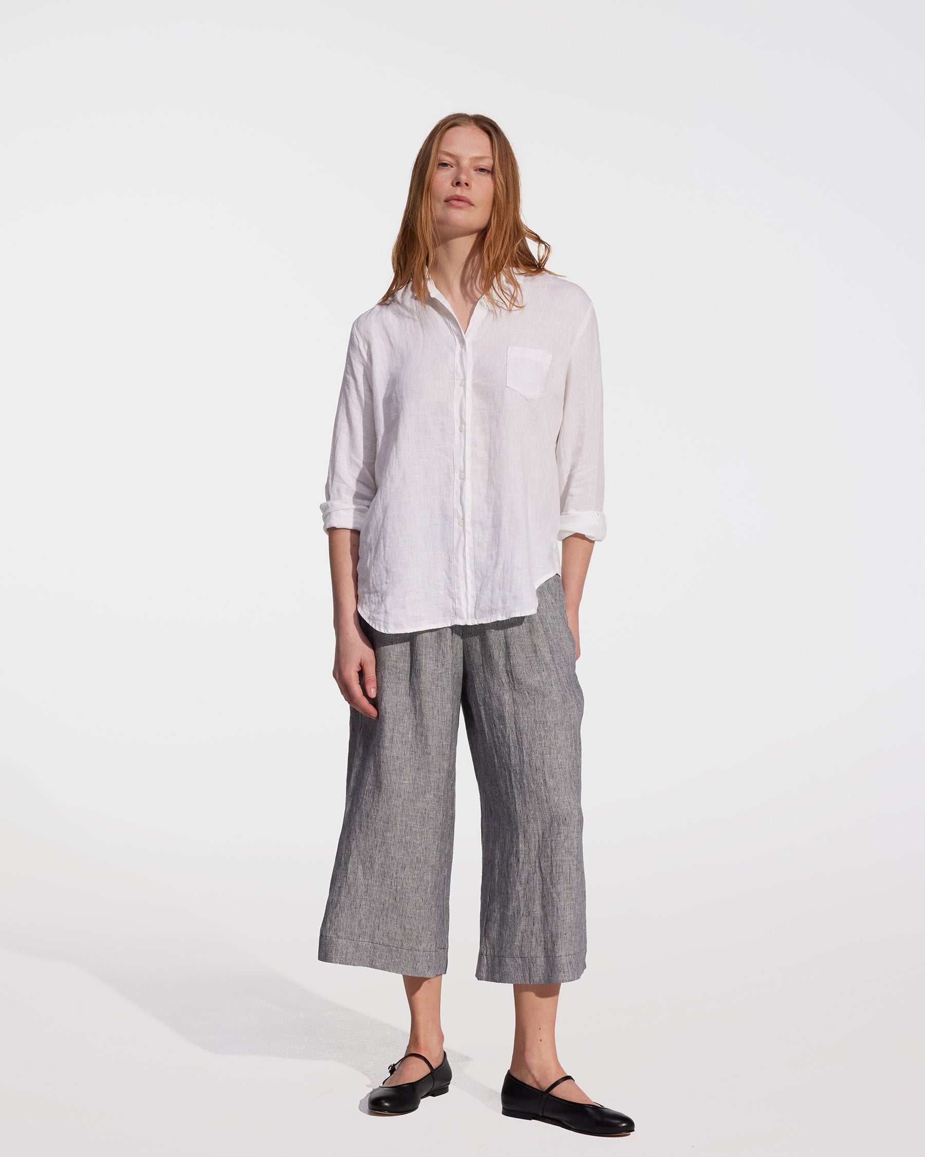 A person stands against a plain white background wearing a white long-sleeve button-up shirt, black flat shoes, and Pyne and Smith Clothiers’ Linen Culottes No.11 in Cooke's Stripe, with one hand in their pocket.