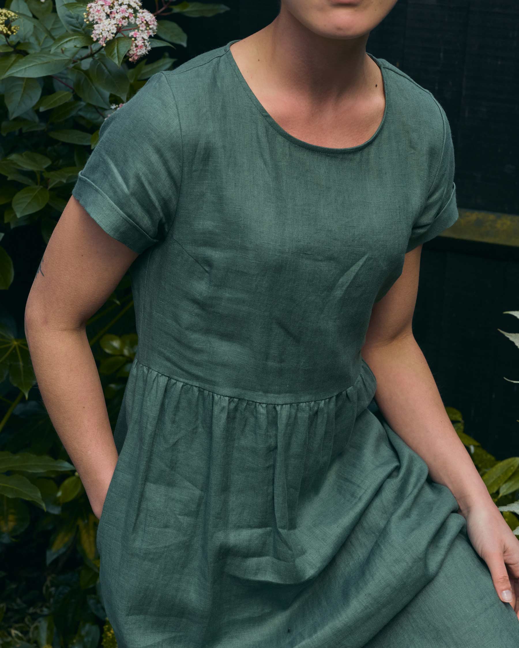 A person wearing the No.17 Full Length Linen Tea Dress in Carriage Green by Pyne and Smith Clothiers, made from European flax linen, sits outdoors near green leafy plants with hands in pockets; their face is partially out of frame.
