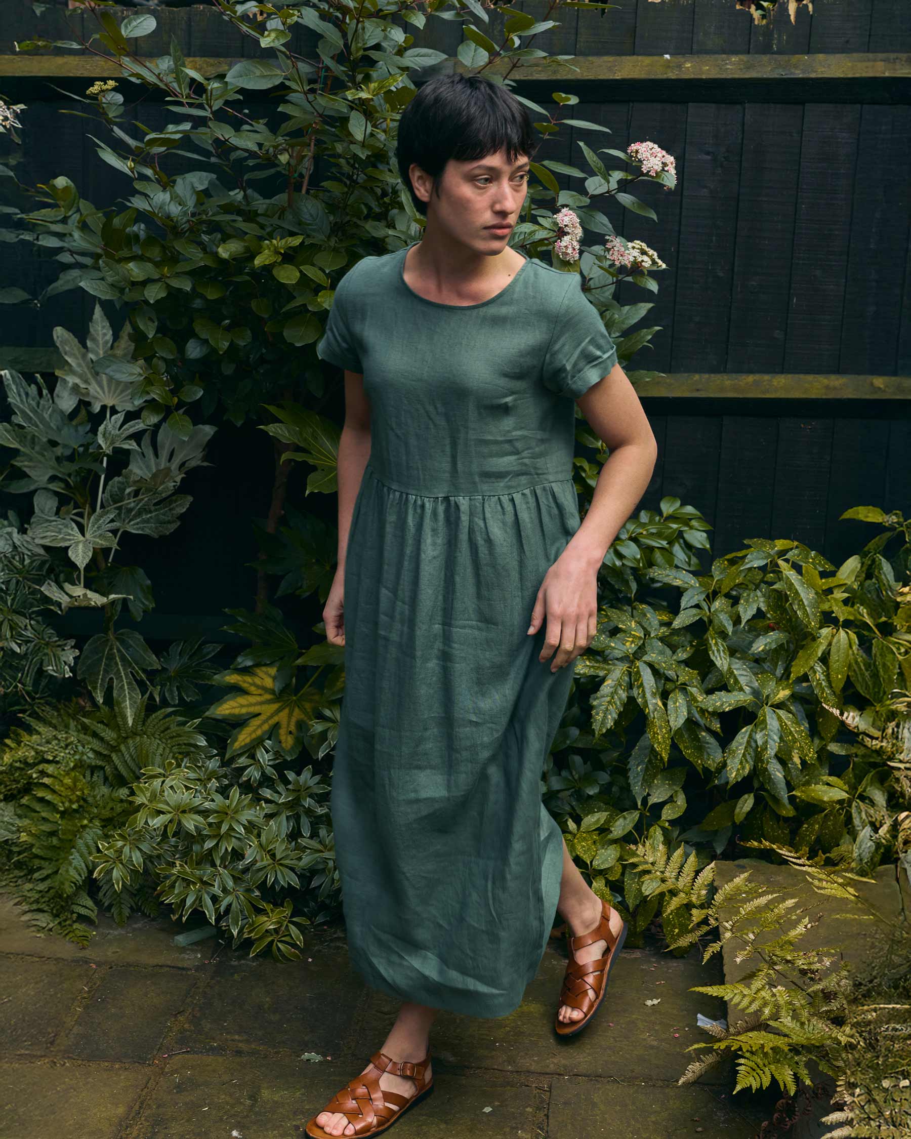 A woman in the Pyne and Smith Clothiers No.17 Full Length Linen Tea Dress in Carriage Green and brown sandals stands outdoors on a stone path, surrounded by lush plants with a dark wooden fence behind her.