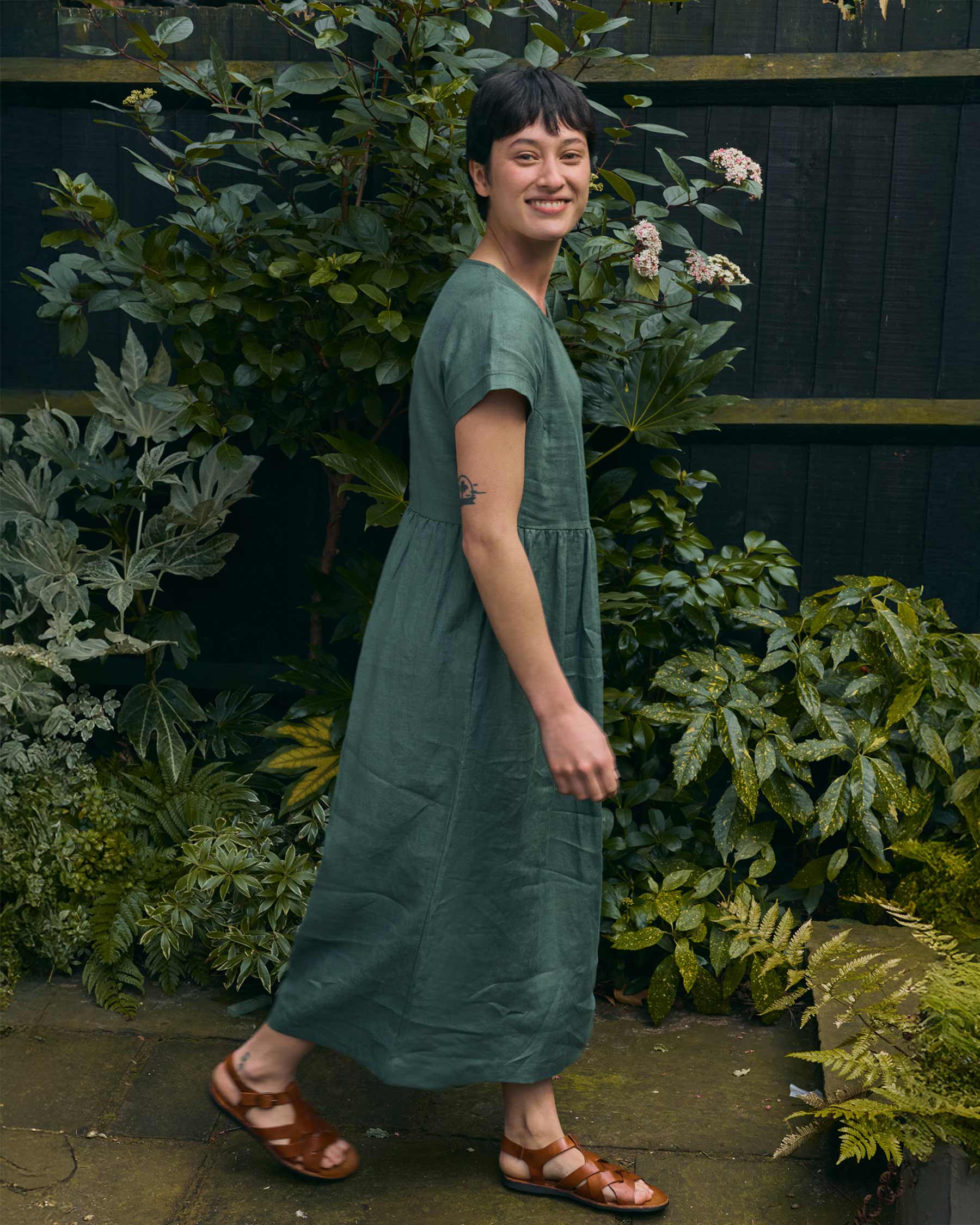 A person with short dark hair smiles while walking outside in the Pyne and Smith Clothiers No.17 Full Length Linen Tea Dress in Carriage Green, paired with brown sandals, surrounded by leafy green plants and a dark wooden fence.