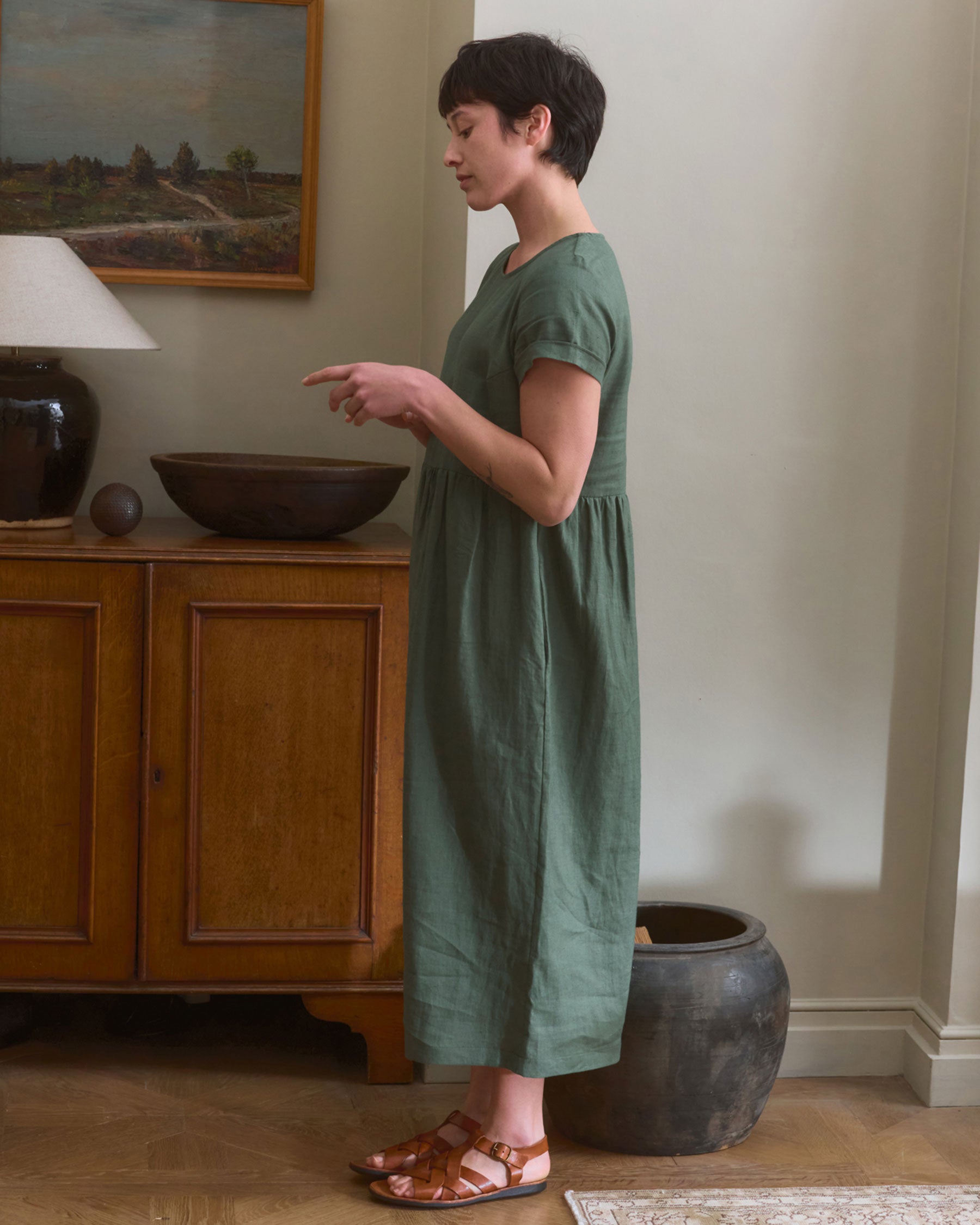 A woman wears the Pyne and Smith Clothiers No.17 Full Length Linen Tea Dress in Carriage Green with brown sandals, standing sideways in a living room beside a wooden sideboard topped with a lamp, bowl, and wall art above.