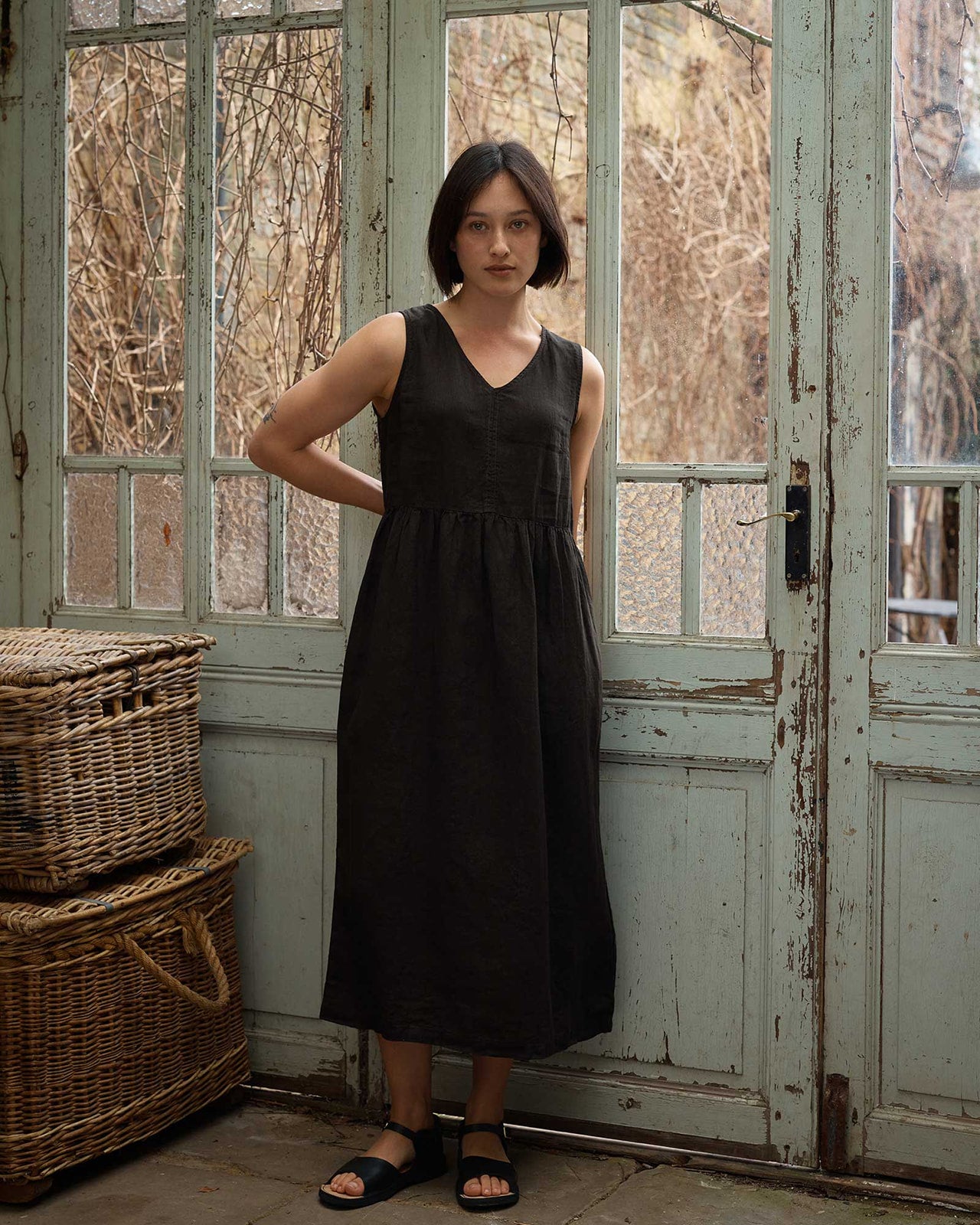 A woman with short dark hair stands indoors by weathered glass doors, wearing a Size Medium #676 No.21 V Neck Sleeveless Linen Dress in Black by Pyne and Smith Clothiers, paired with black sandals, beside stacked wicker baskets. Dried branches show through the glass.
