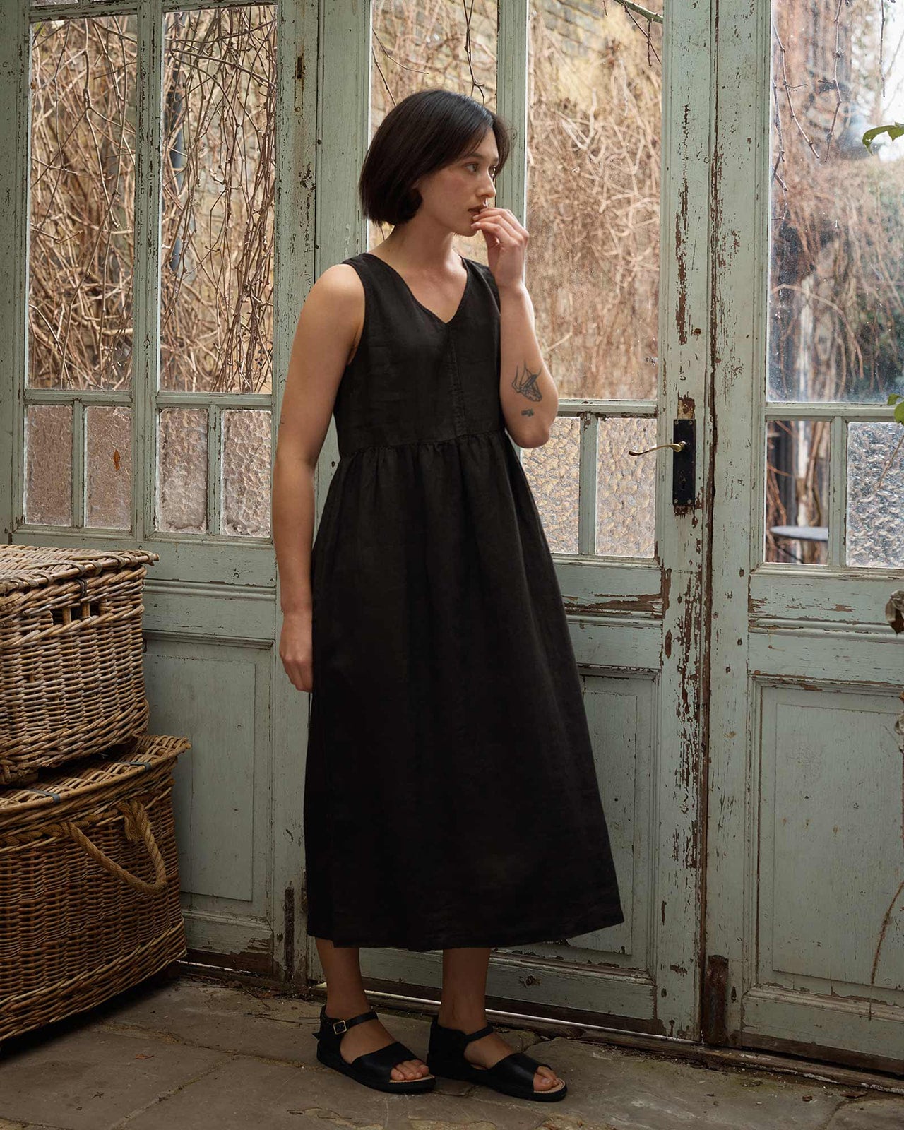 A woman wears the Pyne and Smith Clothiers Size Medium #676 No.21 V Neck Sleeveless Linen Dress in Black, styled with black sandals, standing indoors by large glass doors beside wicker baskets and rustic outdoor branches.