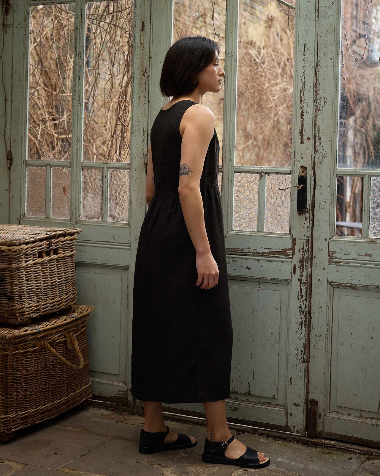 A woman with short dark hair wears the Pyne and Smith Clothiers Size Medium #676 No.21 V Neck Sleeveless Linen Dress in Black, styled with black sandals, standing sideways near tall vintage glass-paned doors beside two stacked wicker baskets.