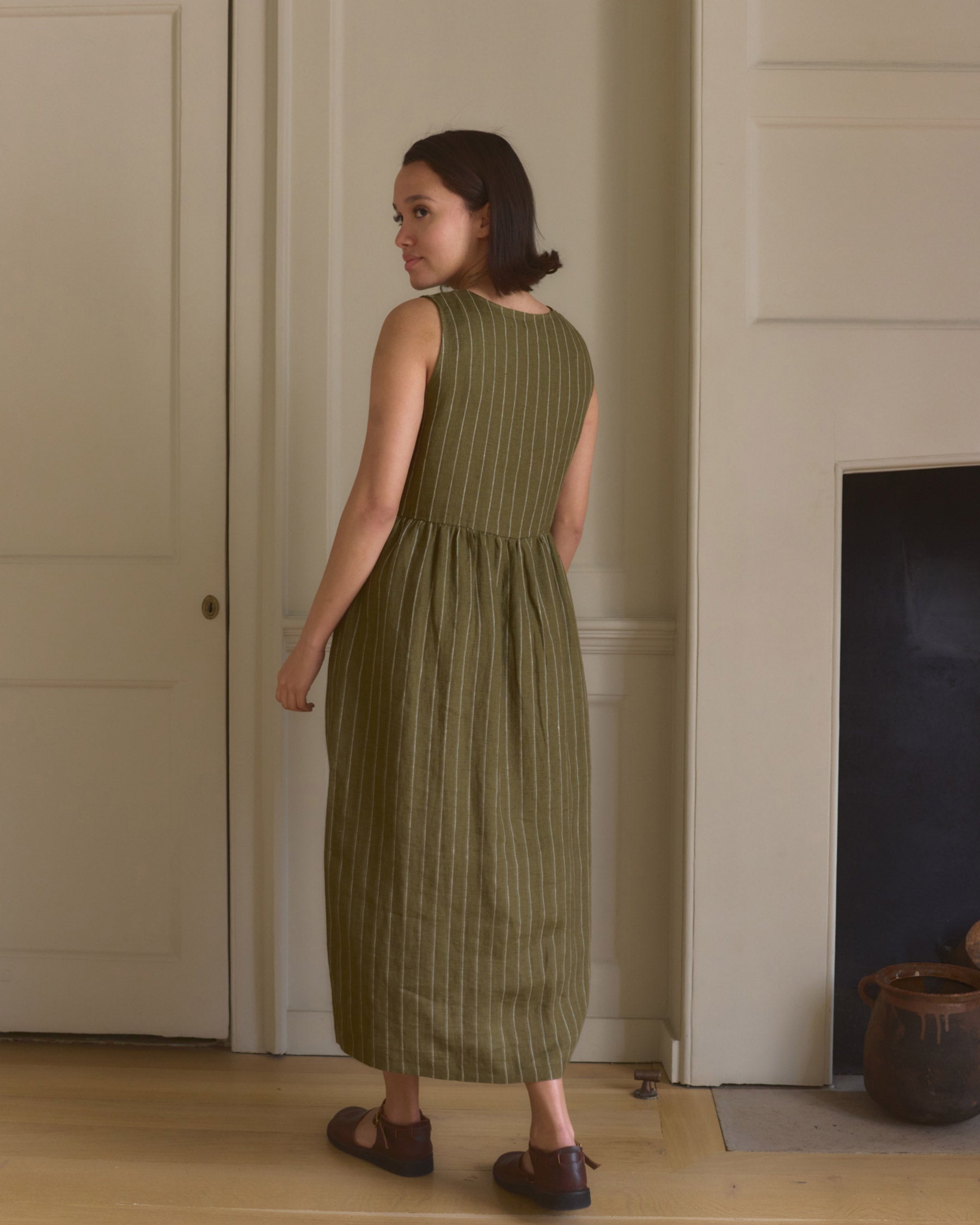 A woman with short brown hair stands indoors by a white wall and fireplace, wearing the Pyne and Smith Clothiers No.21 V Neck Sleeveless Linen Dress in Kelp Green Stripe with brown shoes on a wooden floor.